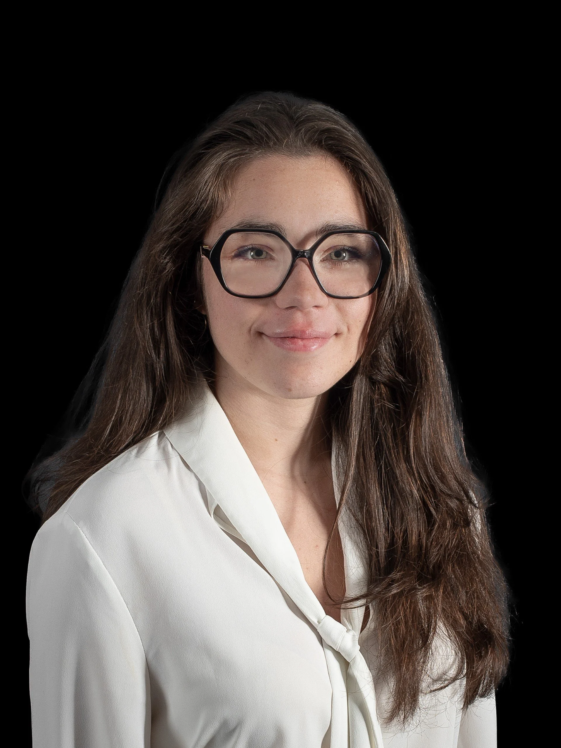 Portrait d'une femme avec de longs cheveux bruns, portant des lunettes épaisses et une chemise blanche, souriant légèrement, fond noir.