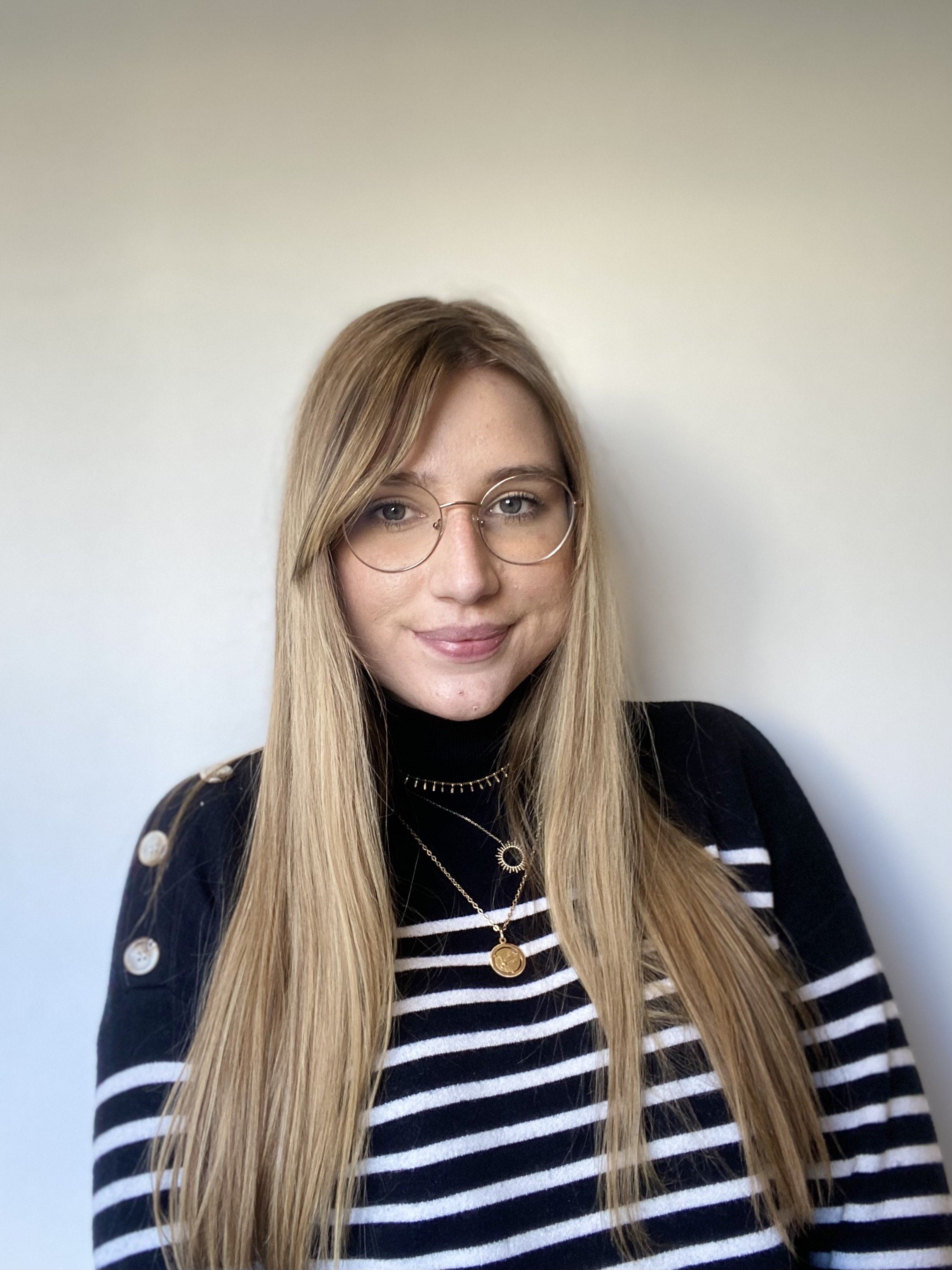 Jeune femme aux longs cheveux blonds portant des lunettes rondes, un pull rayé noir et blanc avec des boutons sur l'épaule, et plusieurs colliers dorés, posant contre un mur blanc.