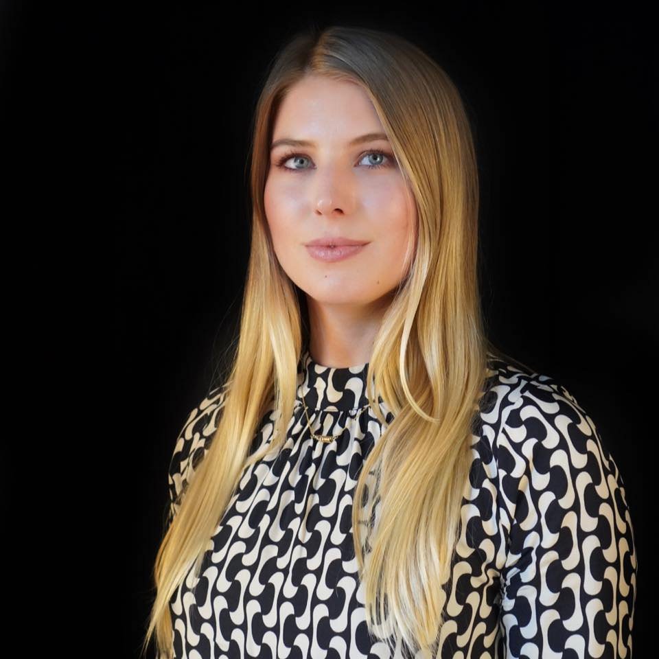 Portrait d'une jeune femme aux longs cheveux blonds, portant une blouse à motifs noirs et blancs, devant un fond noir.