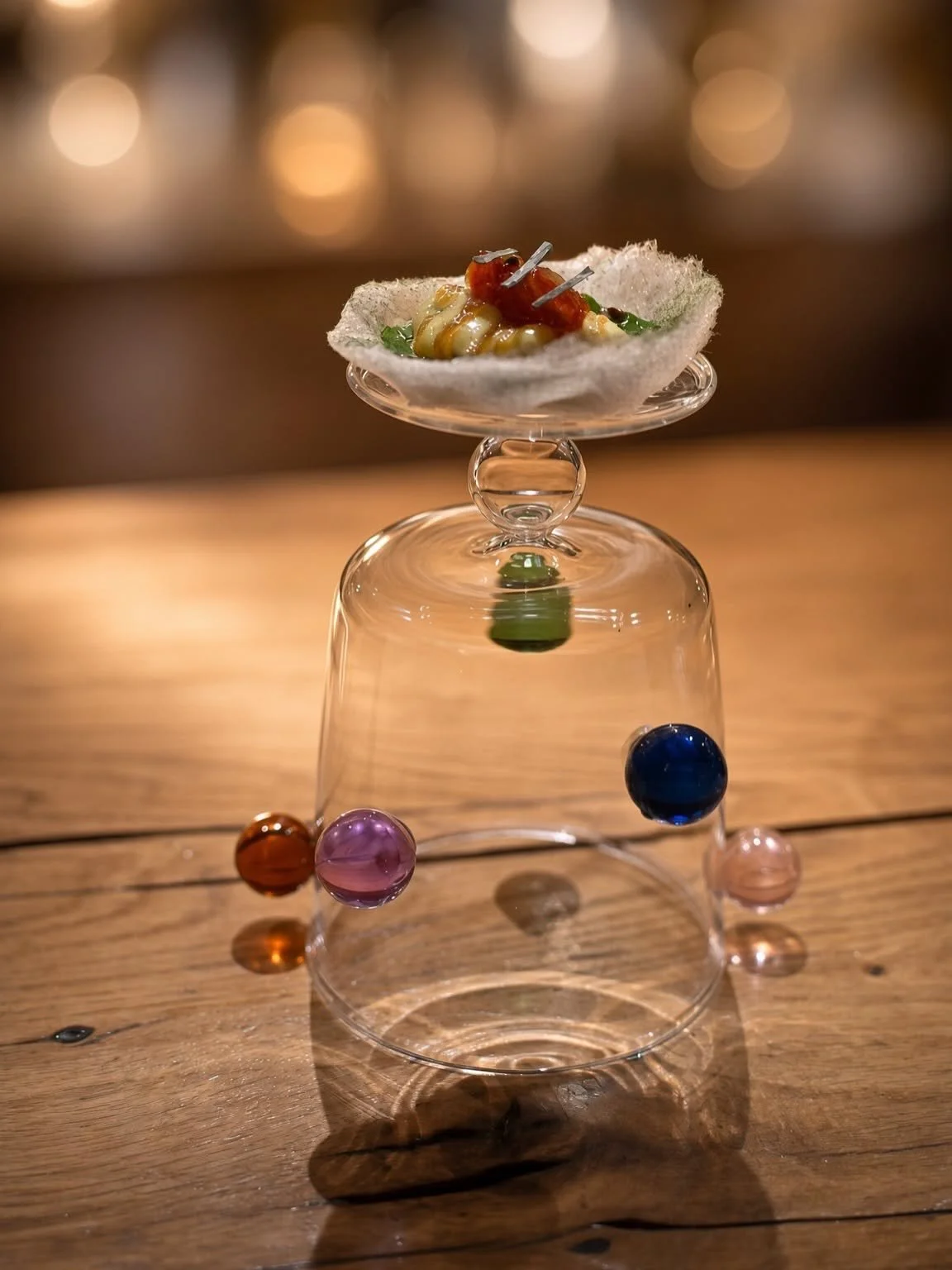 Bouchée présentée au sommet d'une cloche en verre décorée de petites billes colorées, sur le comptoir en bois