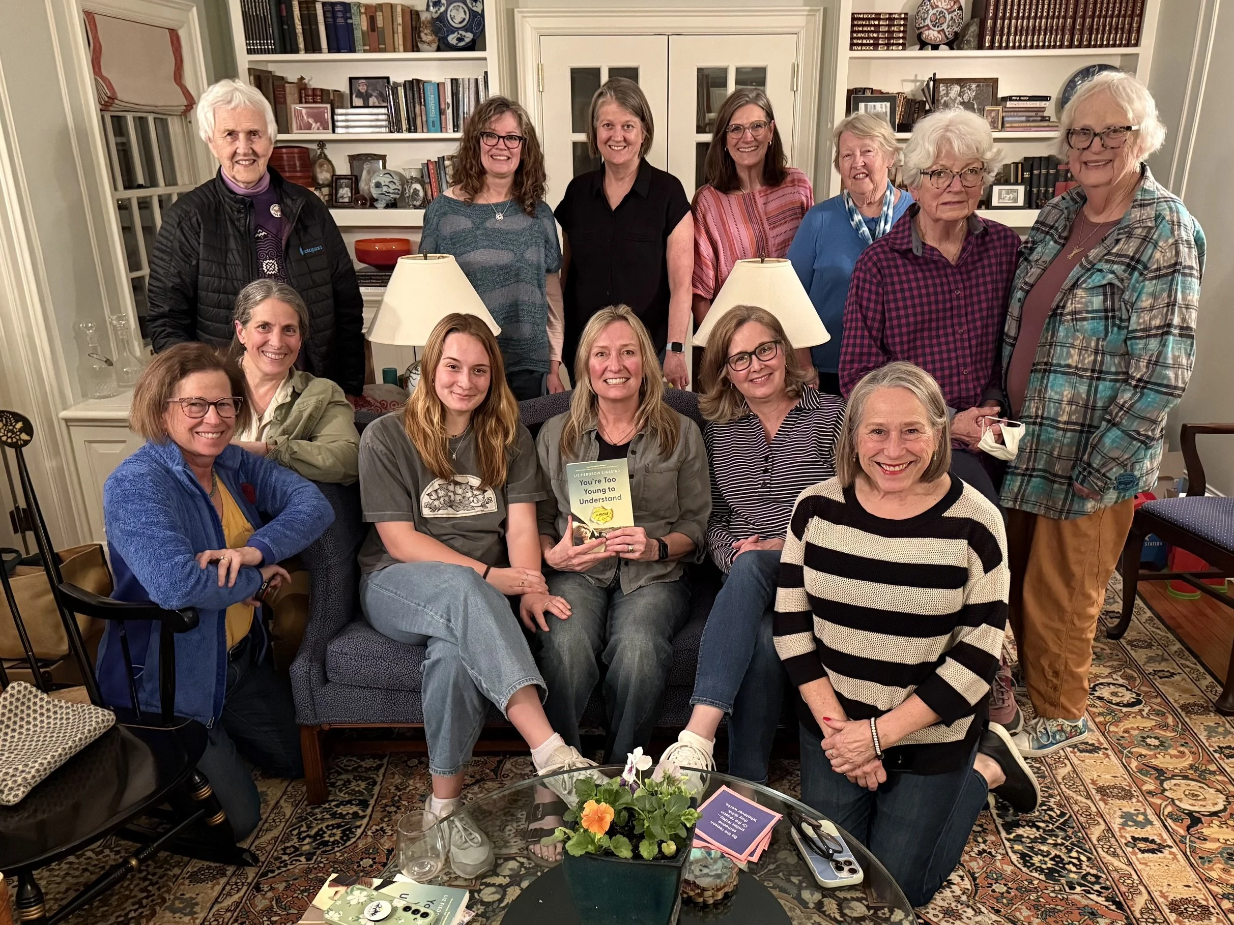 A group of fourteen women, some standing and some sitting, gathered in a warmly lit living room with bookshelves filled with books and decorative items. They are smiling and appear to be celebrating or attending a gathering, with some holding a book titled 'You're Too Young to Understand'