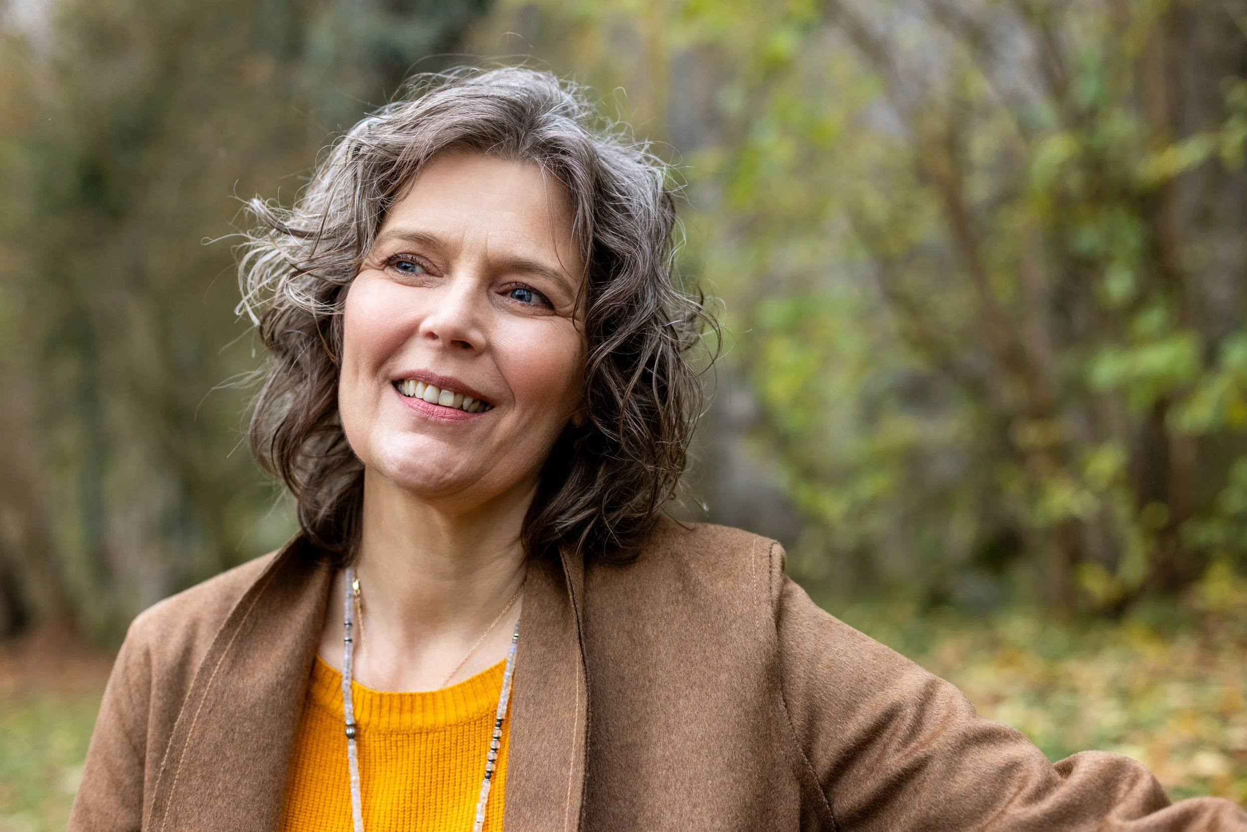 Lächelnde Frau mit lockigem Haar in einem braunen Mantel steht draußen im Grünen.