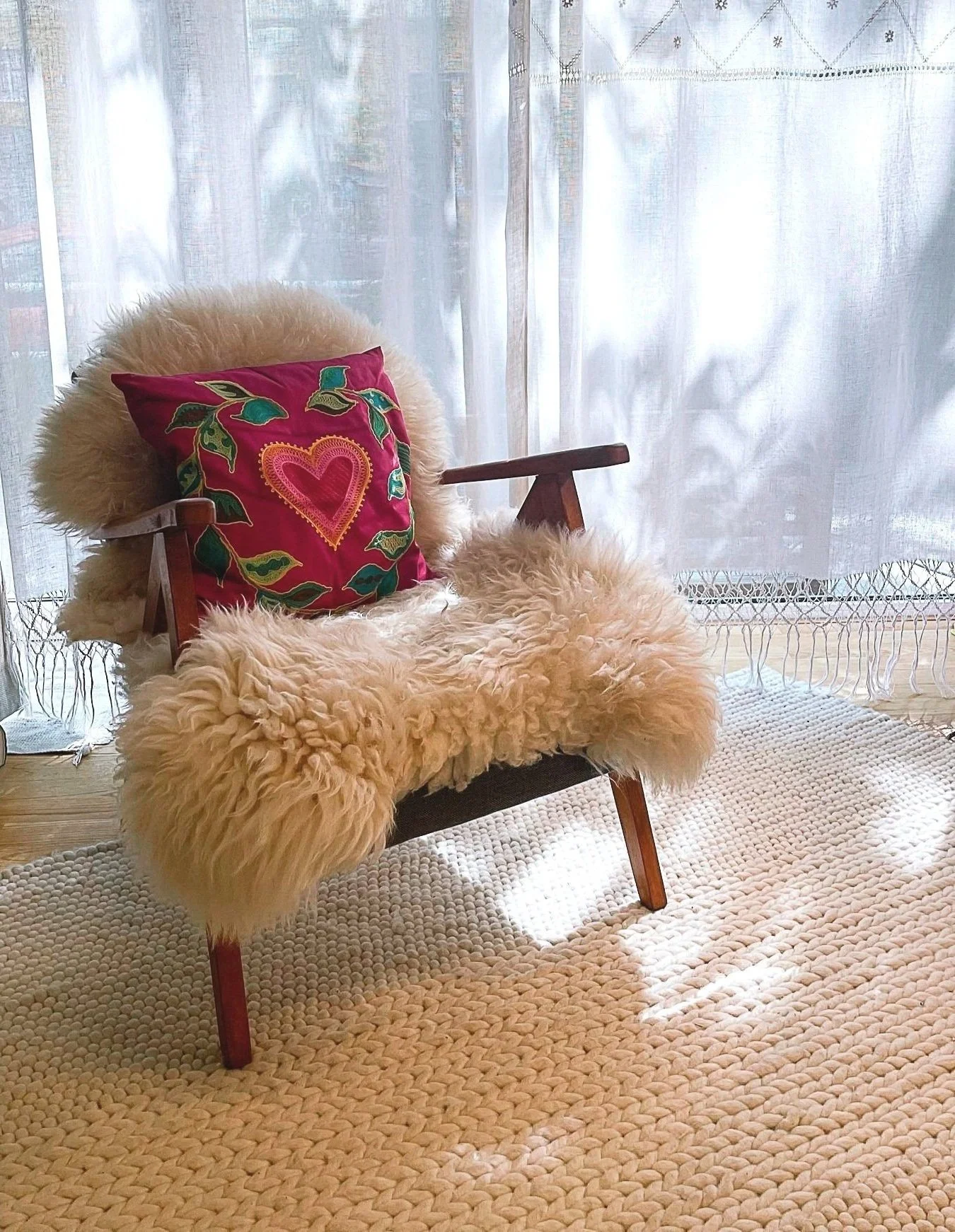 Ein gemütlicher Sessel mit Fellbesatz, eine bunte, bestickte Kissen mit Herz-Design, vor einem Fenster mit halbtransparenten weißen Vorhängen auf einem gerippten, beigen Teppich.