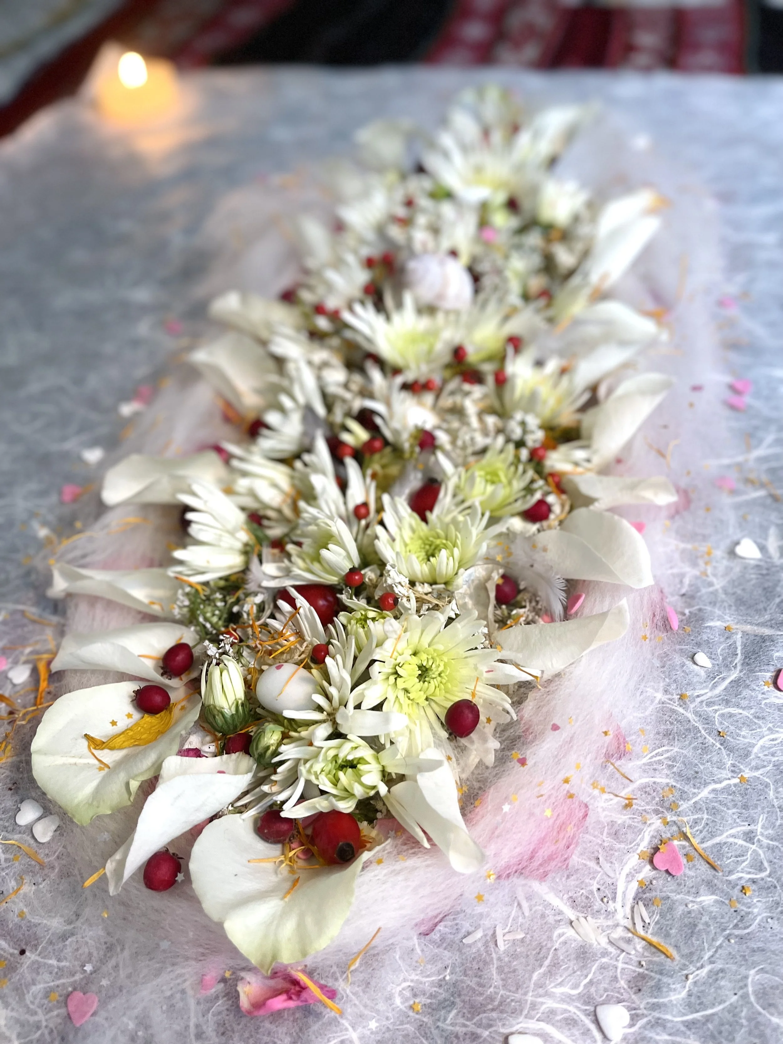 Dekoratives Blumenarrangement mit weißen Blüten, roten Beeren und Blütenblättern auf einer weichen, texturierten Oberfläche, umgeben von verstreuten rosa und goldenen Konfettis. Im Hintergrund brennt eine Kerze.