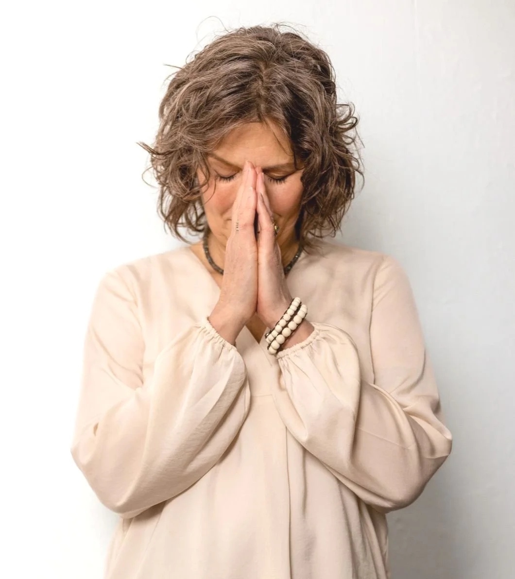 Frau mit geschlossenen Augen und gefalteten Händen, die in Gebet oder Nachdenklichkeit halten, vor hellem Hintergrund.