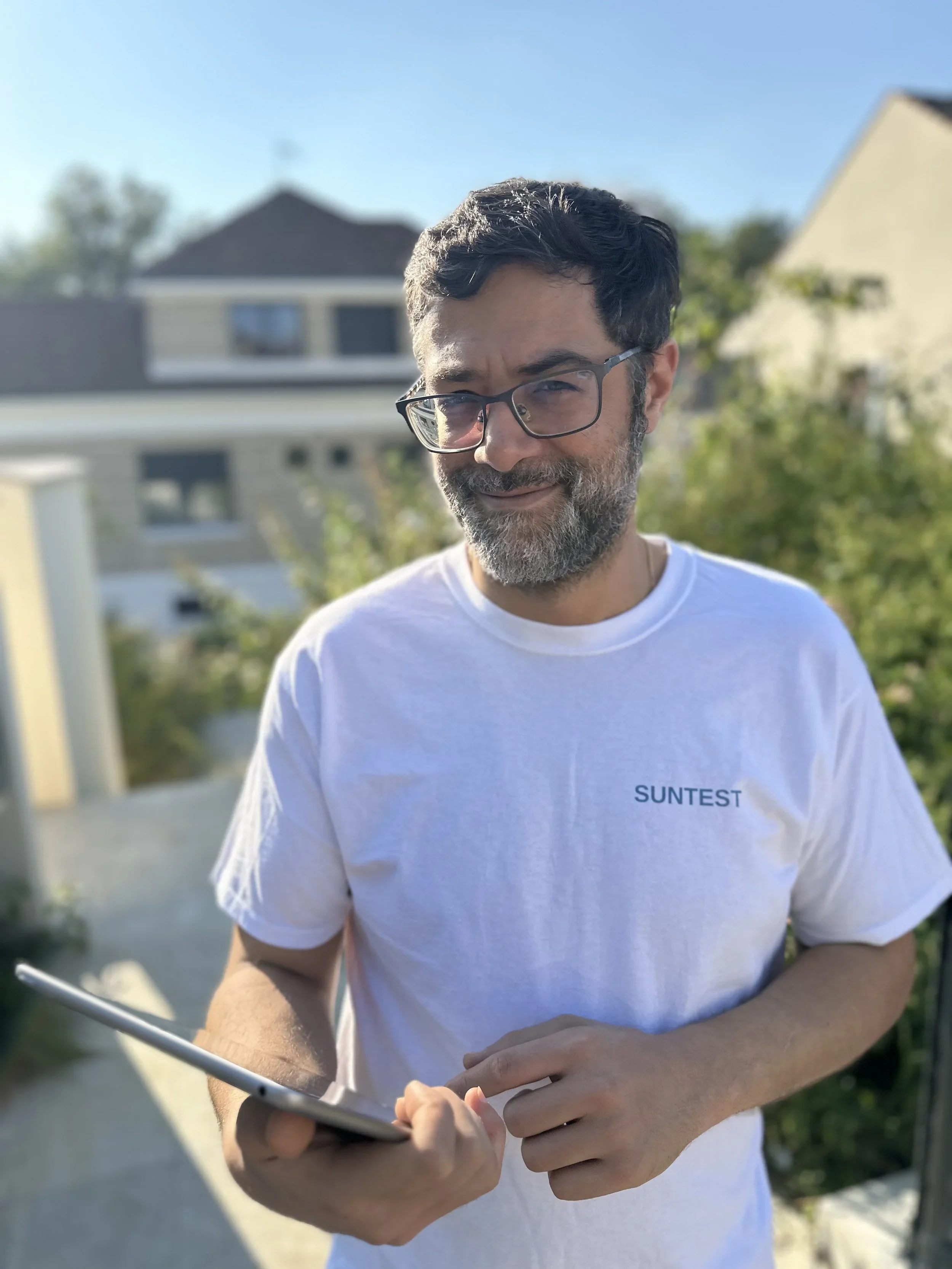 Homme souriant portant des lunettes, tenant une tablette et se trouvant à l'extérieur, avec des maisons et des arbres en arrière-plan par temps clair et ensoleillé.