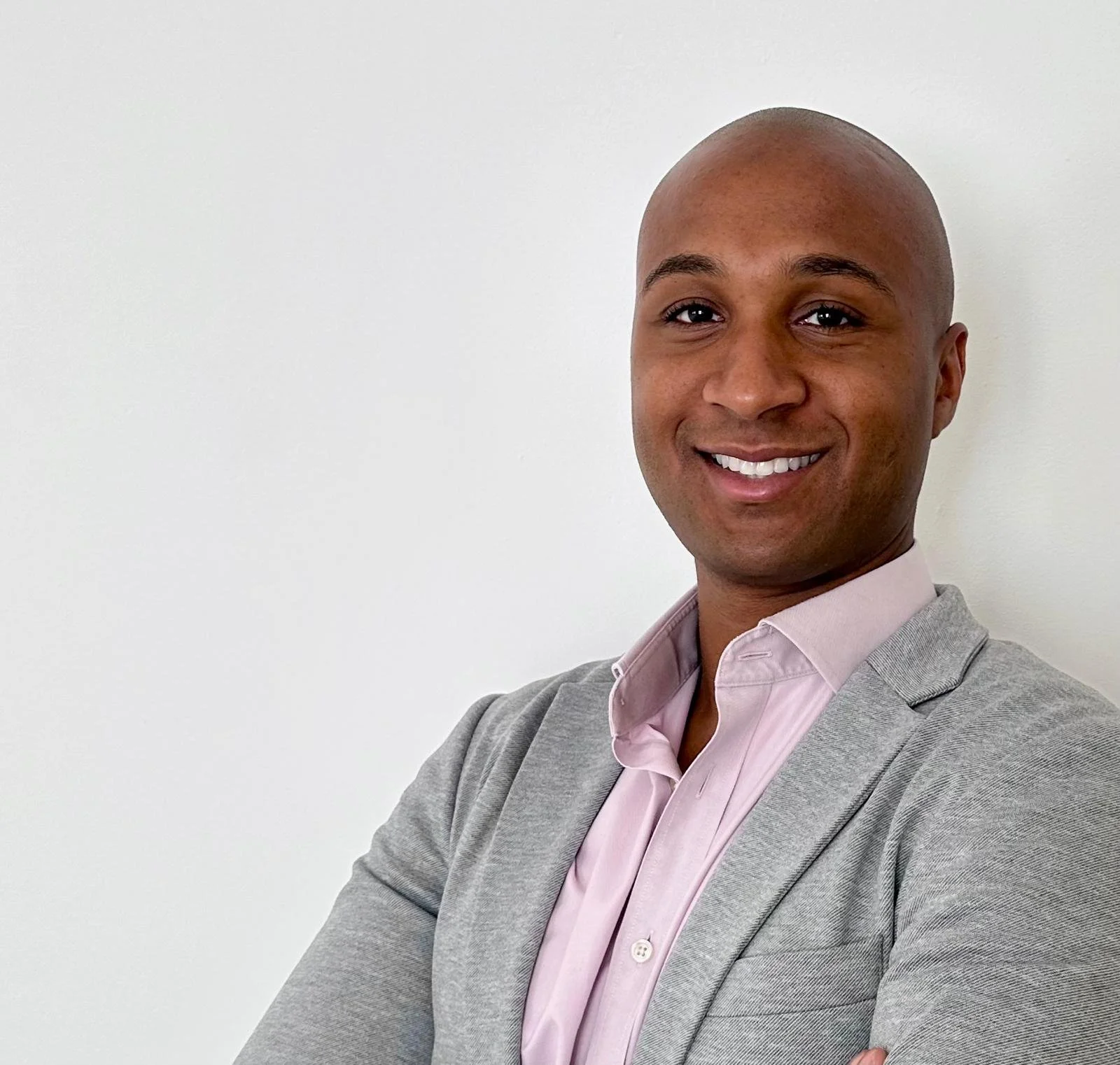 Un homme souriant, portant un blazer gris et une chemise rose clair, se tenant contre un mur blanc.