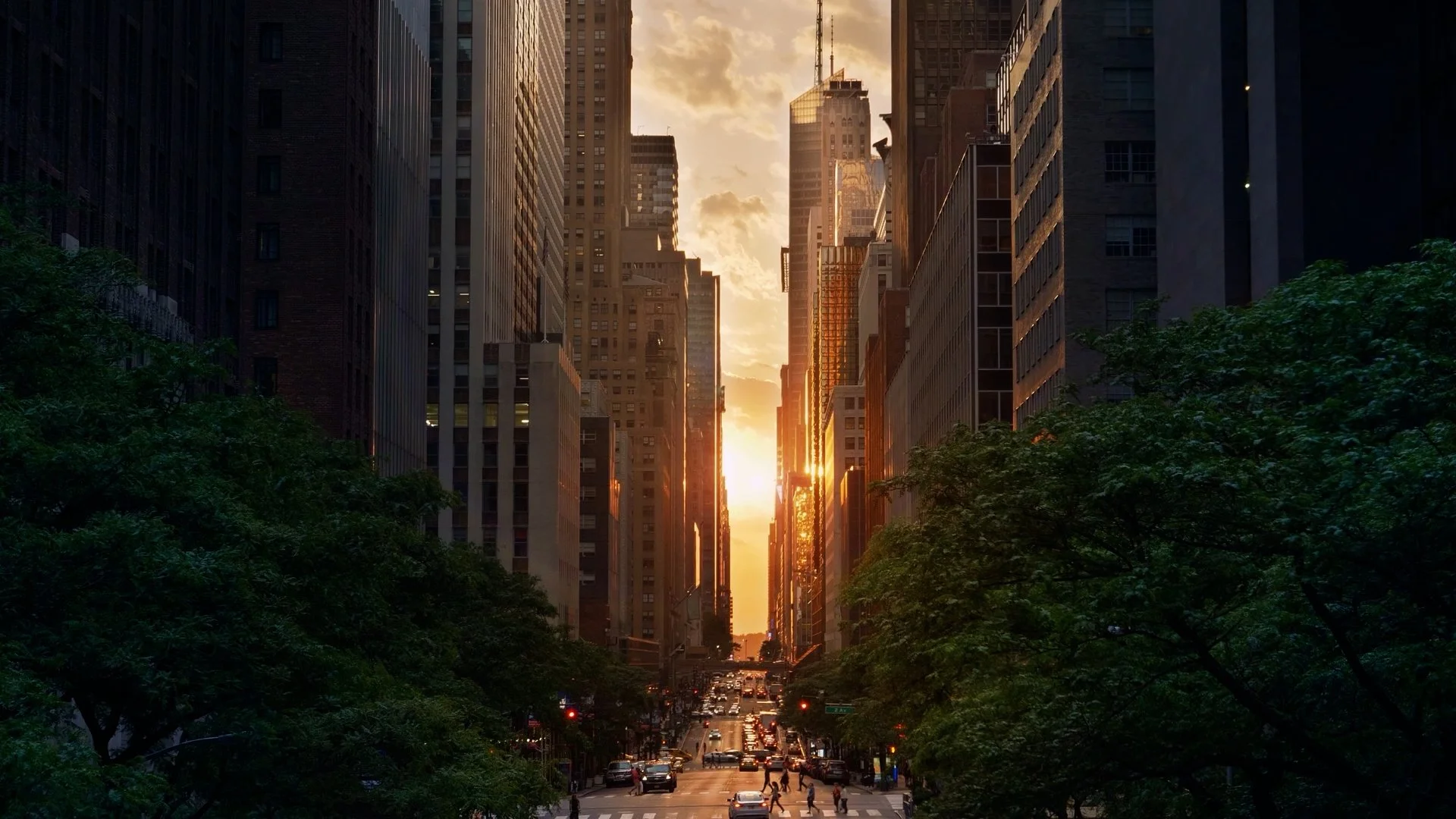 NYC city street at sunset with skyscrapers on both sides and people crossing at the crosswalk.
