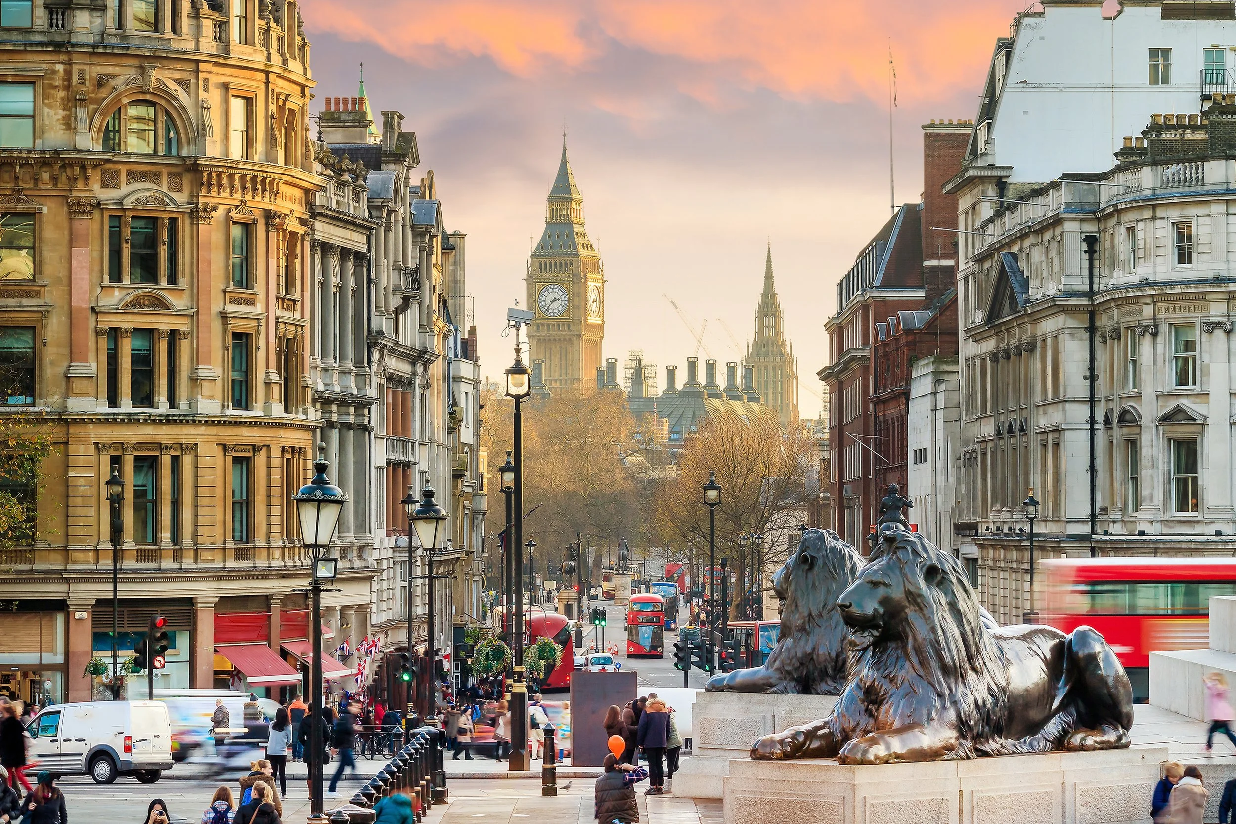 Street scene in London with historic buildings, two lion statues in foreground, Big Ben clock tower in distance, and people walking on the street during sunset.