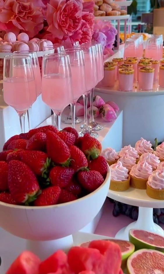 Dessert table with pink drinks in glasses, pink cupcakes, strawberries, macarons, and flower decorations.