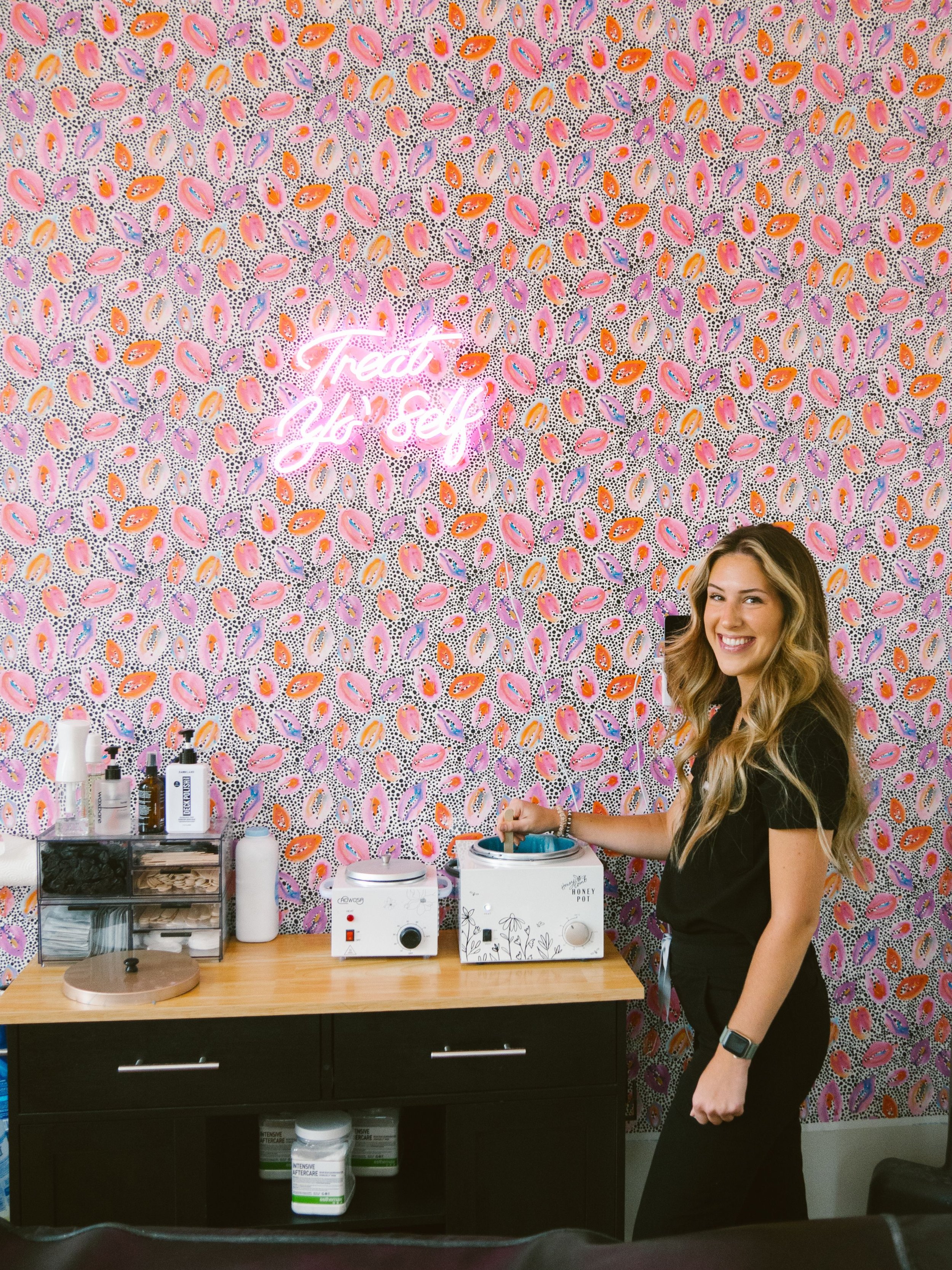 A woman standing next to a wooden countertop with beauty supplies, smiling in front of a colorful, paisley-patterned wall with a neon sign that reads 'Treat Yo' Self.'