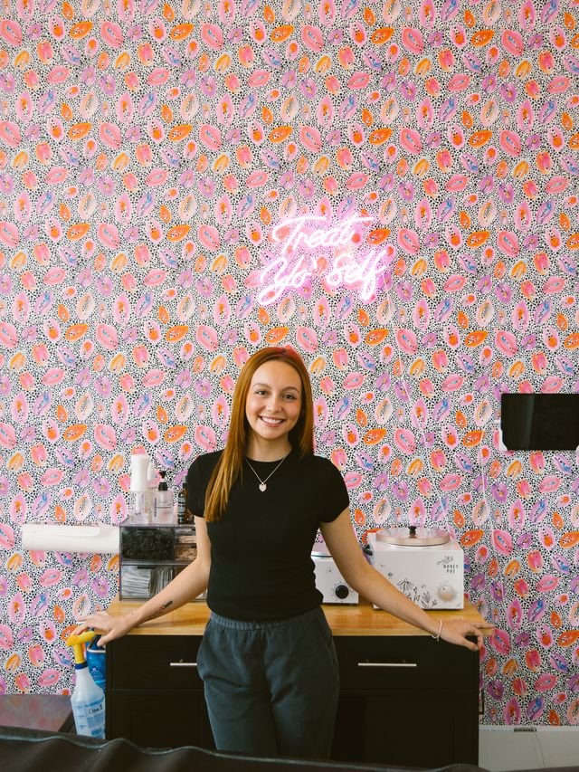 Person standing in front of a colorful patterned wall with a neon sign saying "Treat Yo' Self". They are in a room with a countertop holding beauty or spa equipment, including a wax warmer and cleaning supplies.