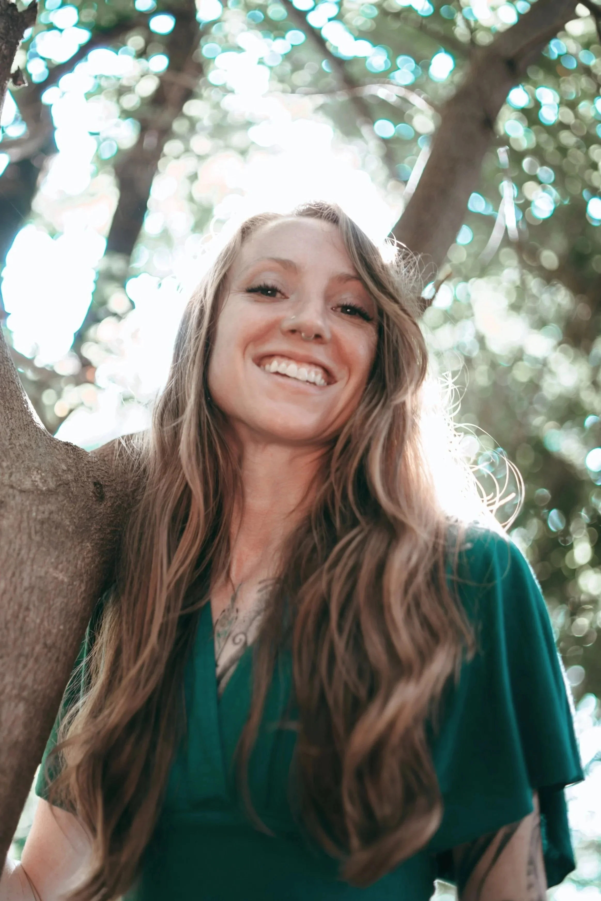 A woman with long wavy hair smiling outdoors near a tree with sunlight filtering through the leaves. Astrologer and Herbalist, Ciera Sims.