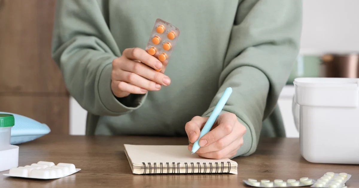 A person has a notepad on a table in front of them. They are holding pills in one hand and writing something down with the other.