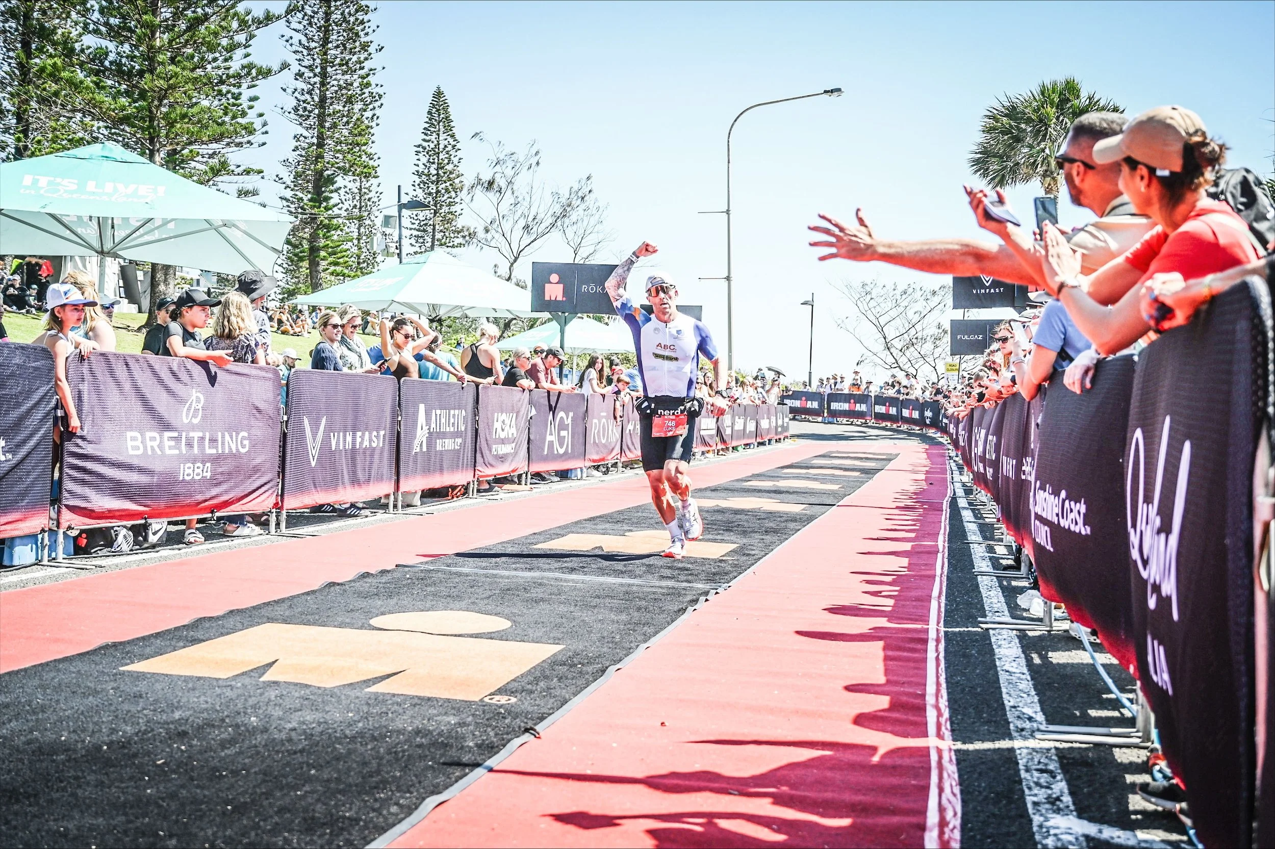 Triathlete celebrating near finish line, surrounded by cheering spectators. Ironman branding visible on ground and barriers. Sunny outdoor setting with trees and umbrellas.