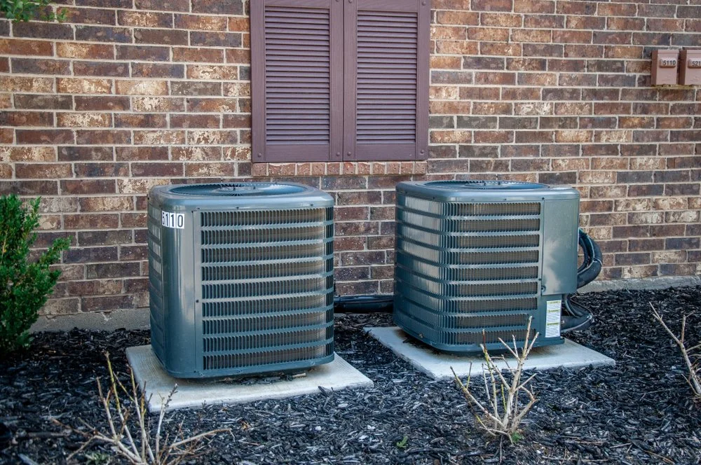 Two outdoor HVAC units next to a brick wall