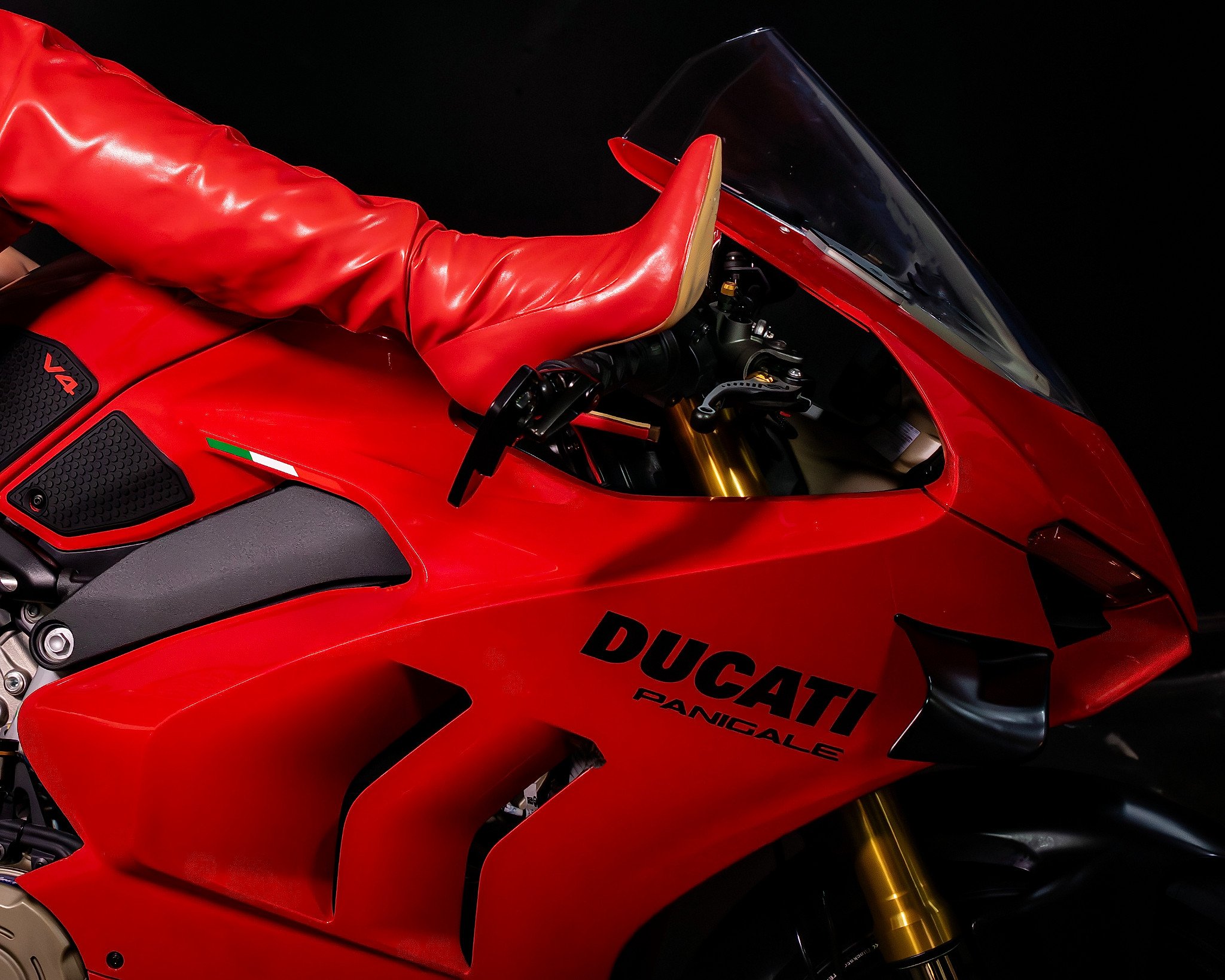 Close-up of a red Ducati Panigale V4 motorcycle with a red seat, black windshield, and gold front fork against a black background.