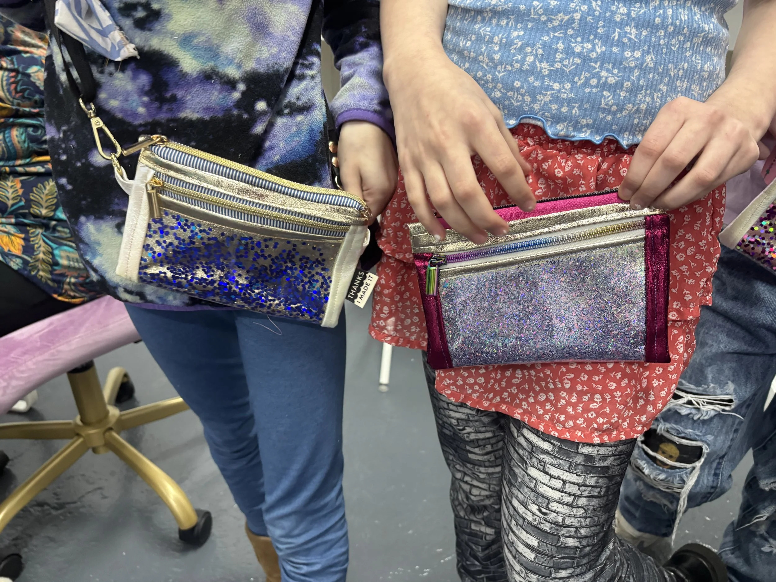 Two people standing side by side holding glittery pouches with zippers, wearing colorful and patterned clothing.