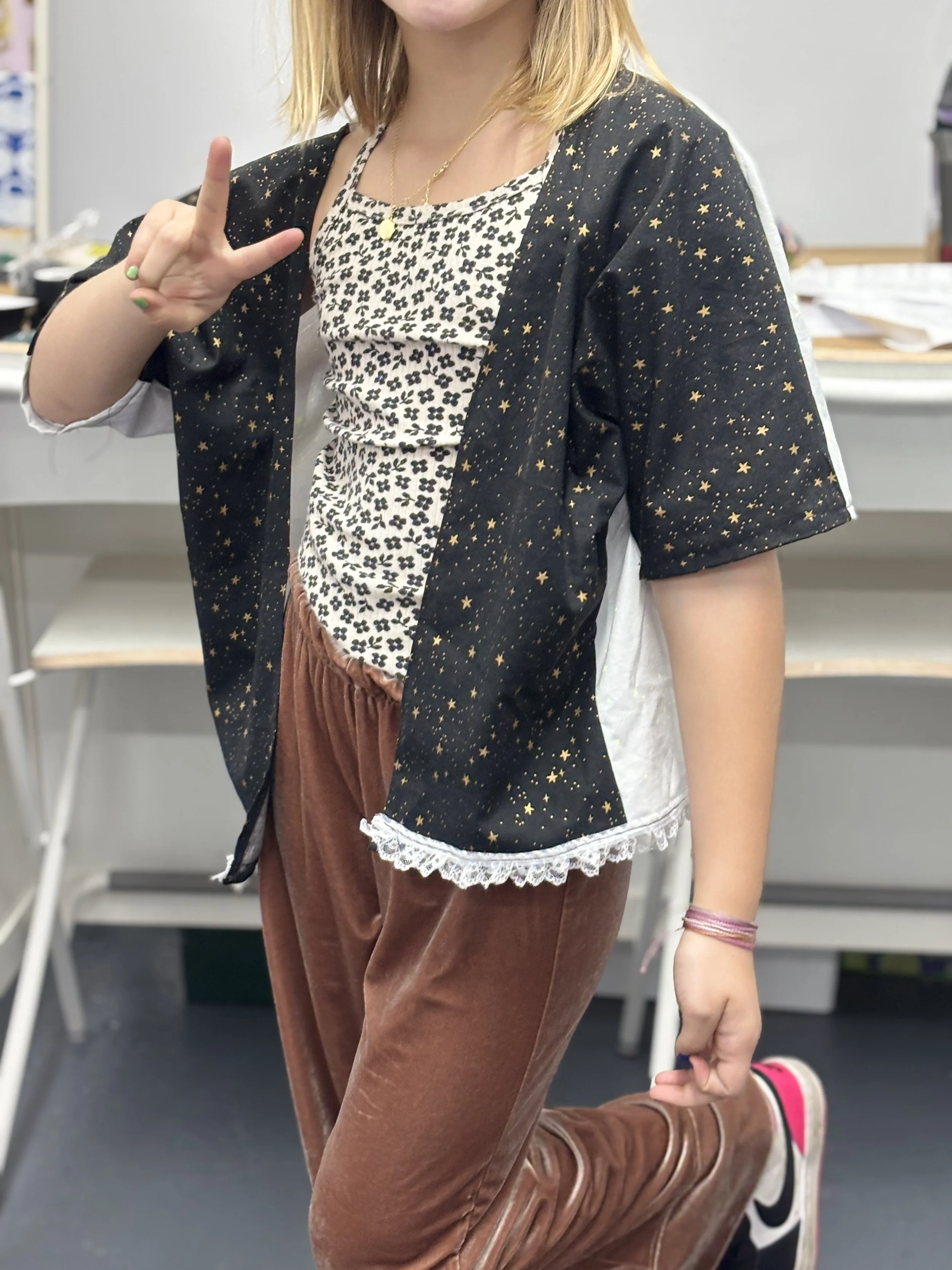 A person with blonde hair making a peace sign with their fingers, dressed in a star-patterned black jacket over a patterned shirt, brown velvety pants, and sneakers, standing in an indoor setting with a desk and papers in the background.