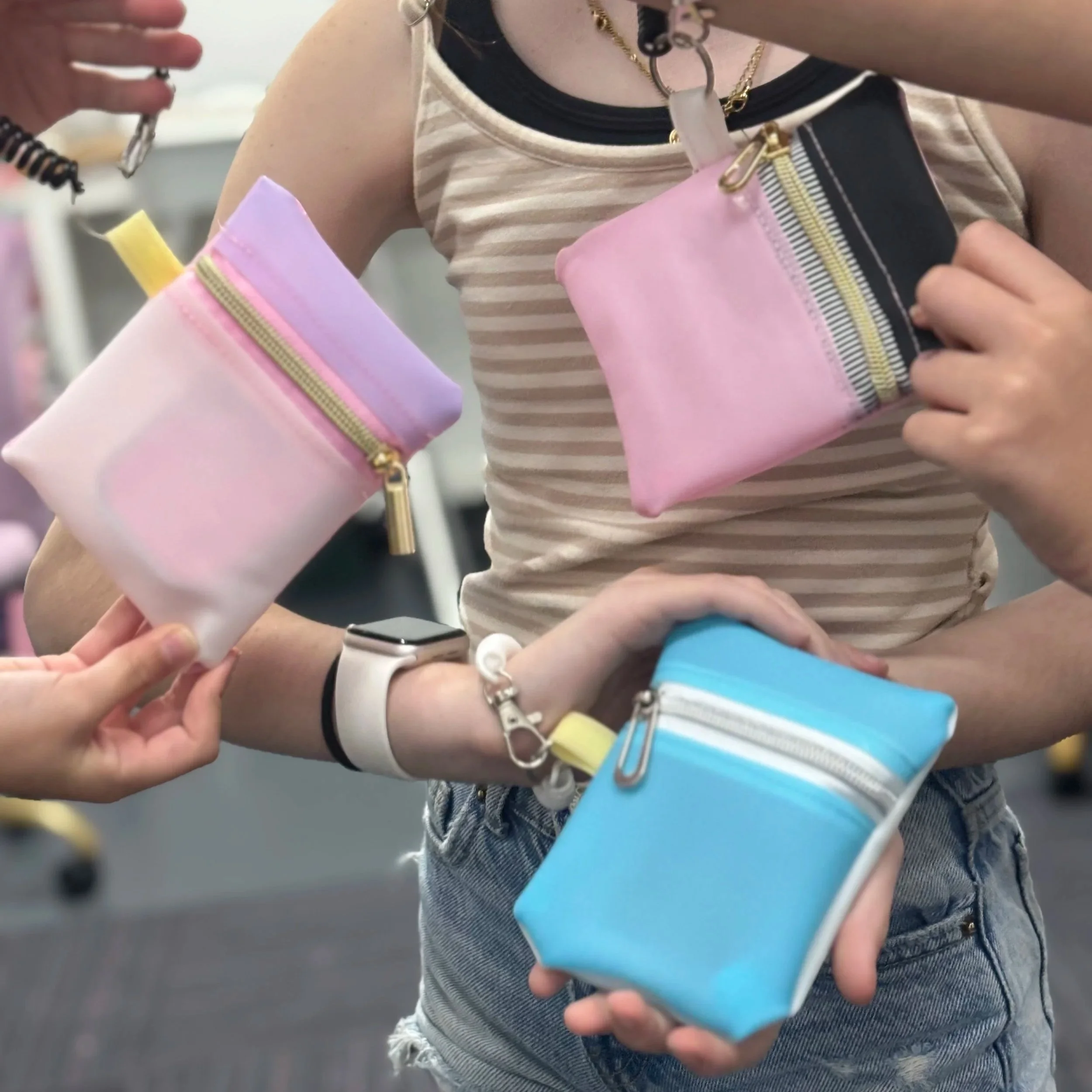 Person holding pastel-colored small pouches with zippers in pink, blue, purple, and black and white, among others. The person is wearing a sleeveless striped top, a watch, and layered necklaces and is in a setting that appears to be an indoor public space.