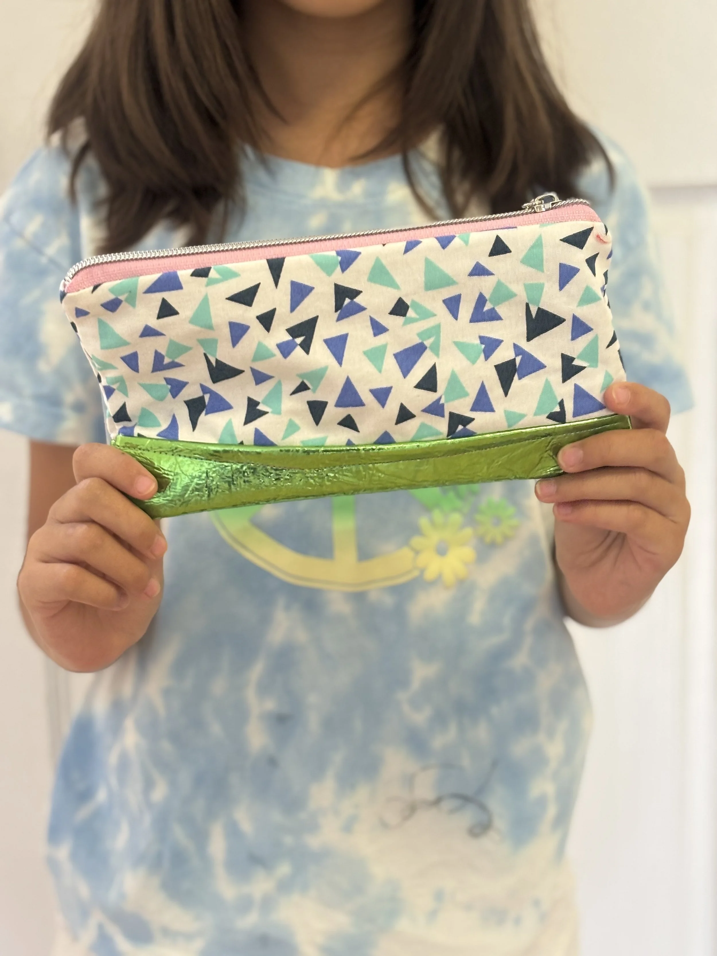 A person holding a colorful patterned clutch bag, wearing a blue tie-dye t-shirt with a peace sign and flower design.