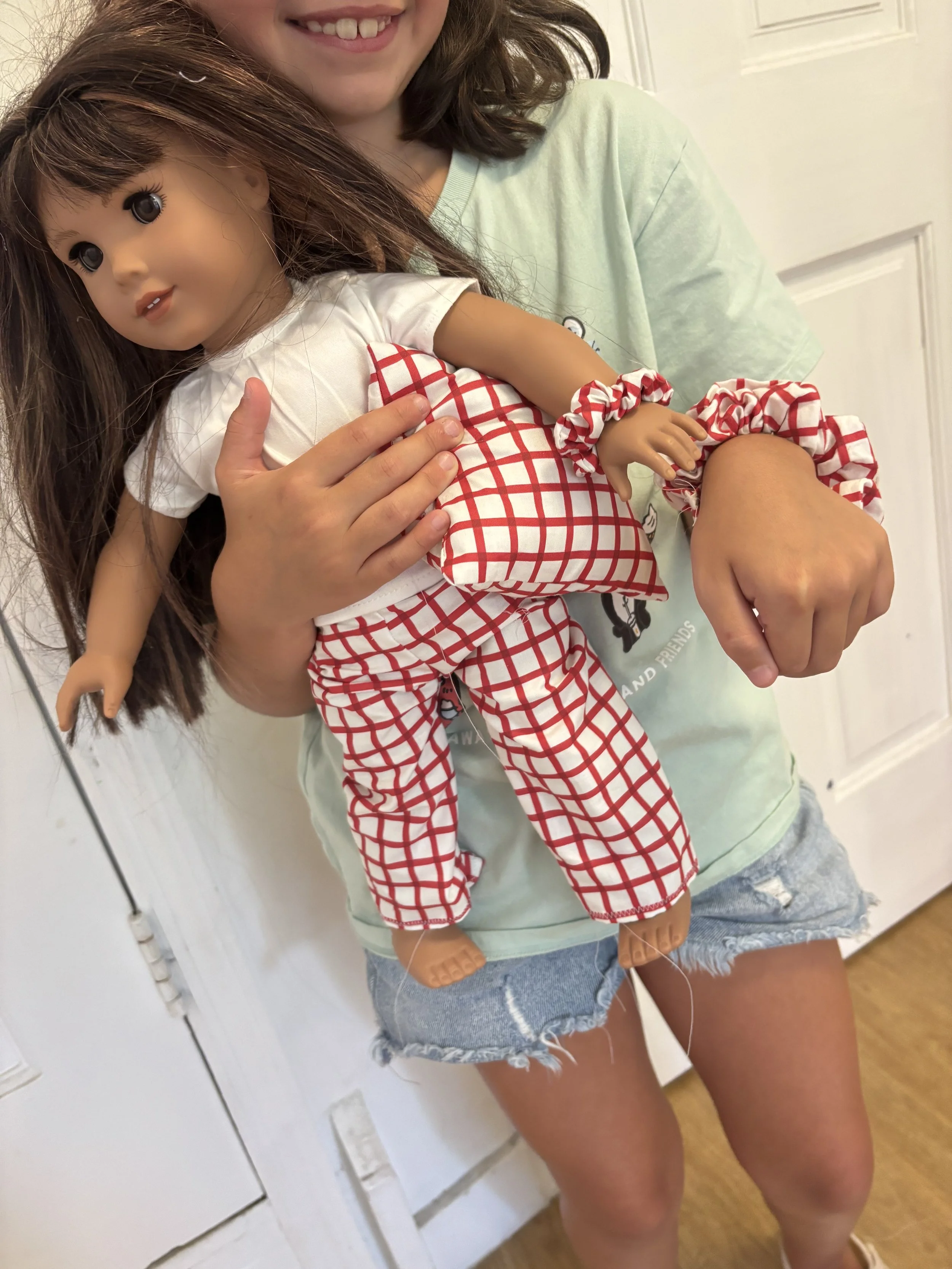 Person holding a doll with brown hair, wearing a red and white checkered outfit, in a room with white walls and wooden floor.