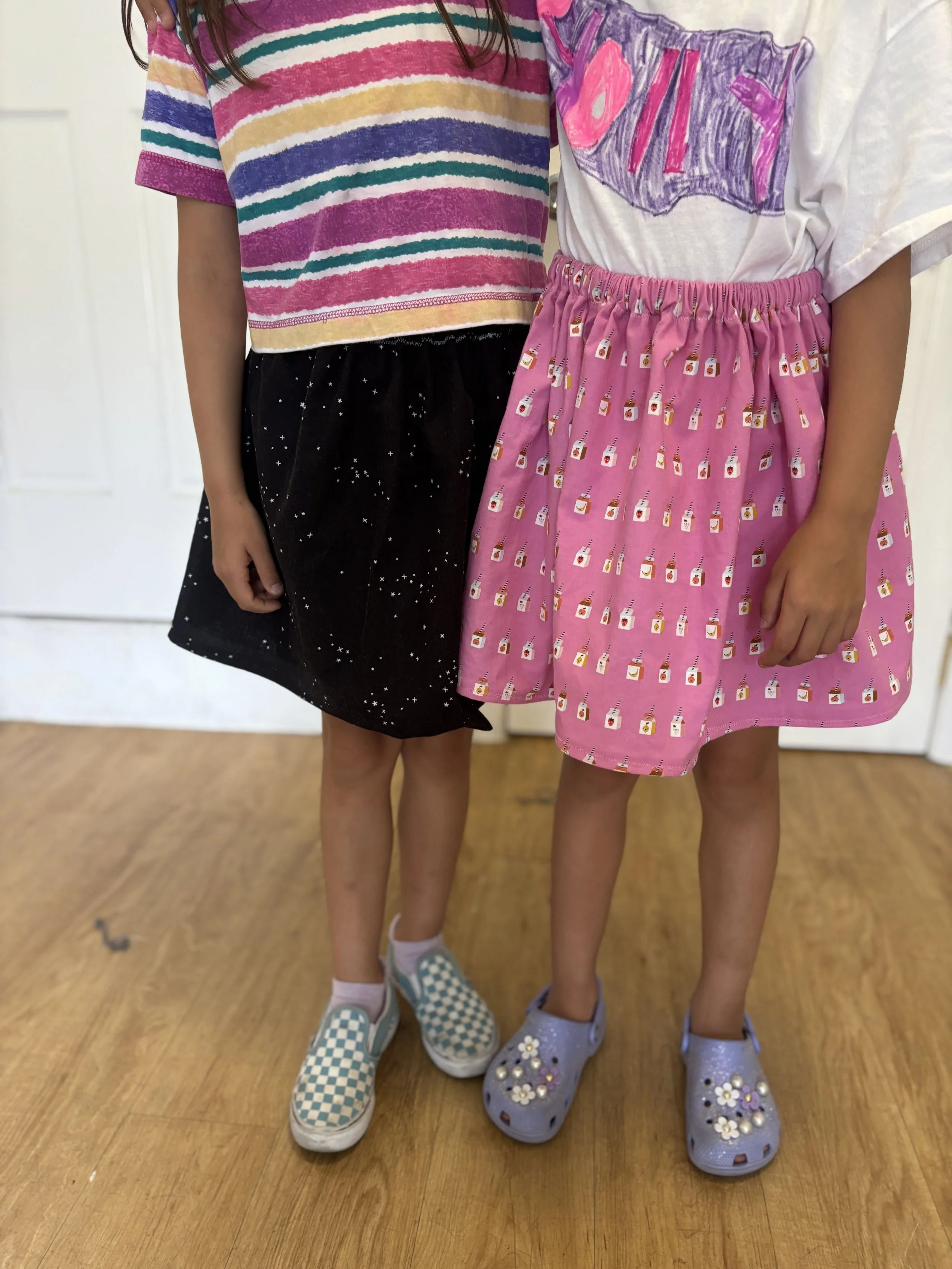 Two children standing on a wooden floor with white door and wall in the background. One child is wearing a striped multicolor t-shirt, a black skirt with white stars, and checkered slip-on shoes. The other child is wearing a white t-shirt with purple and pink lettering, a pink skirt with small milk carton patterns, and purple Crocs decorated with flowers.