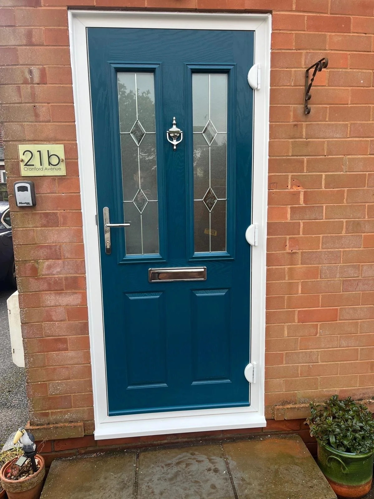 Blue front door with glass panels, mail slot, door knocker, and door handle, flanked by a brick wall, with house number 21b on a sign to the left, a doorbell, and potted plants nearby.