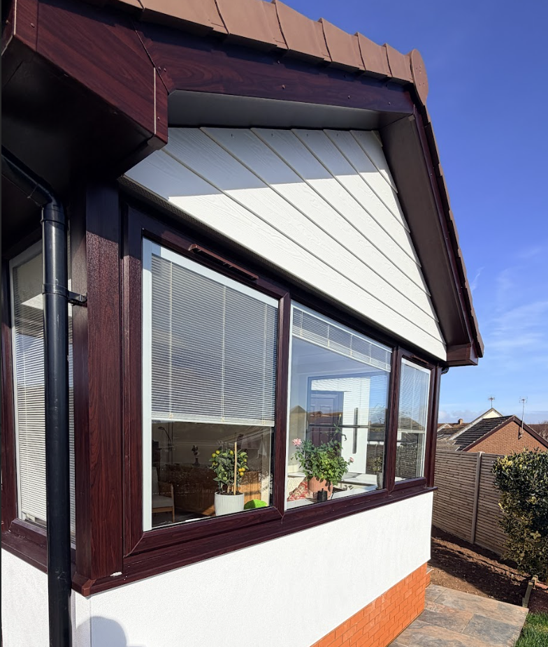 Close-up of a house corner with brown wooden framing, glass windows with blinds, and a white and red brick exterior, under a clear blue sky.
