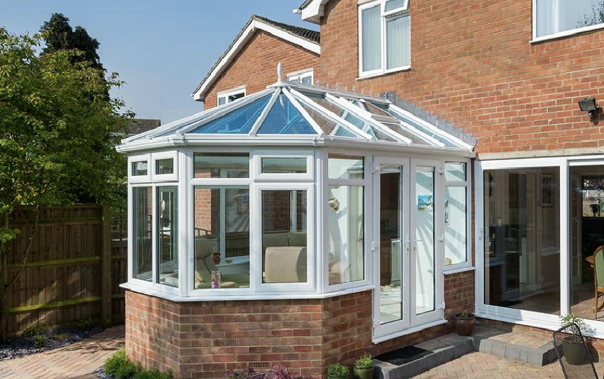 A brick house with a glass conservatory or sunroom attached to the back, featuring large windows and a glass roof, with some indoor furniture visible.