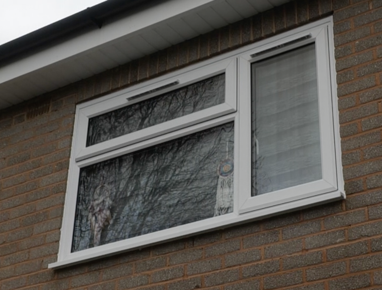 Exterior view of a brick house window with three panes, one top horizontal window and two large lower windows, reflecting trees and sky, with white window frames and a hanging decoration inside.