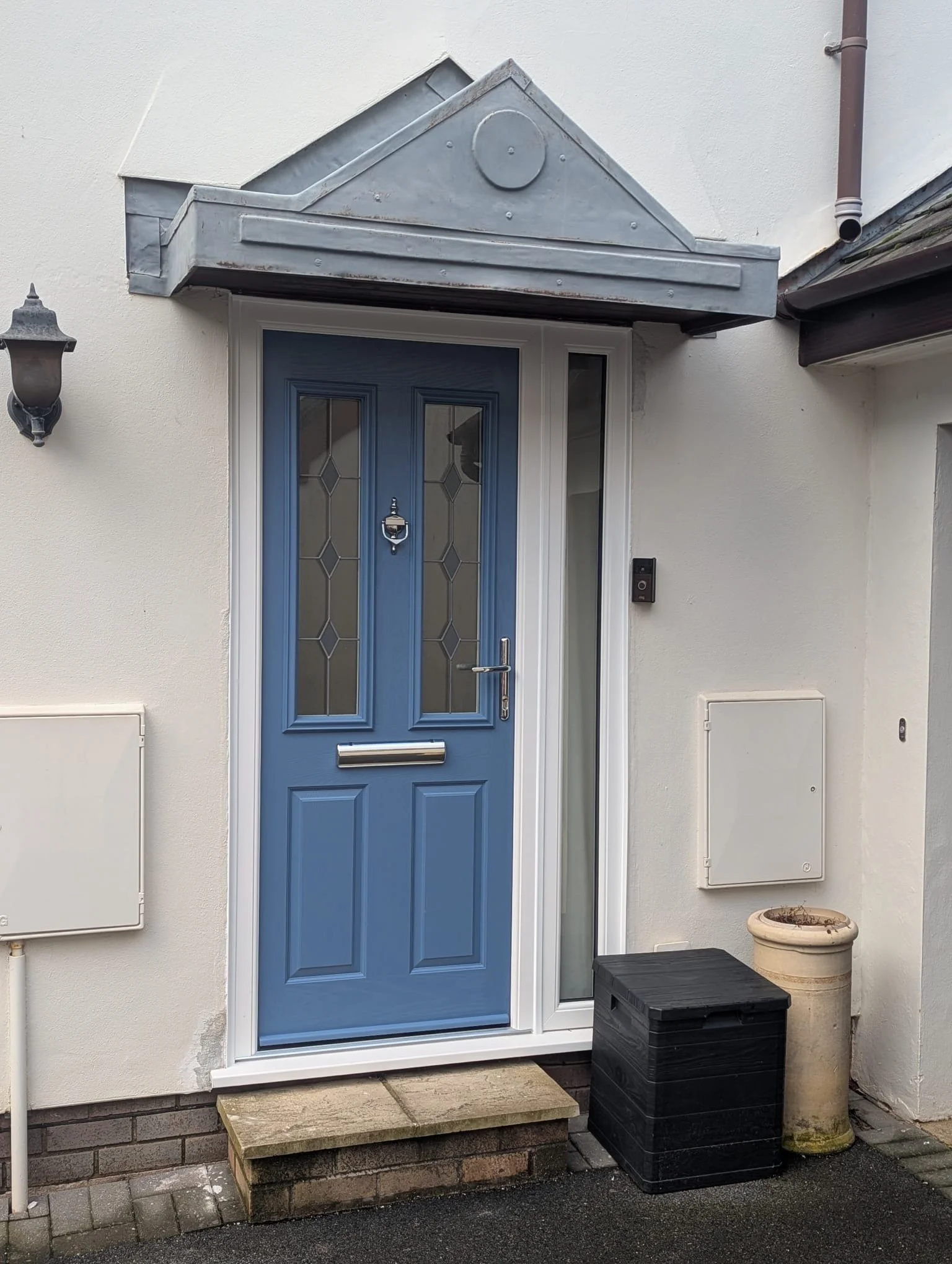 Residential front entrance with a blue door featuring glass panels, a mail slot, a door knocker, and a handle. The door is framed by white walls, with a small step leading up to it. To the right, there is a black storage box and an old flower pot. To