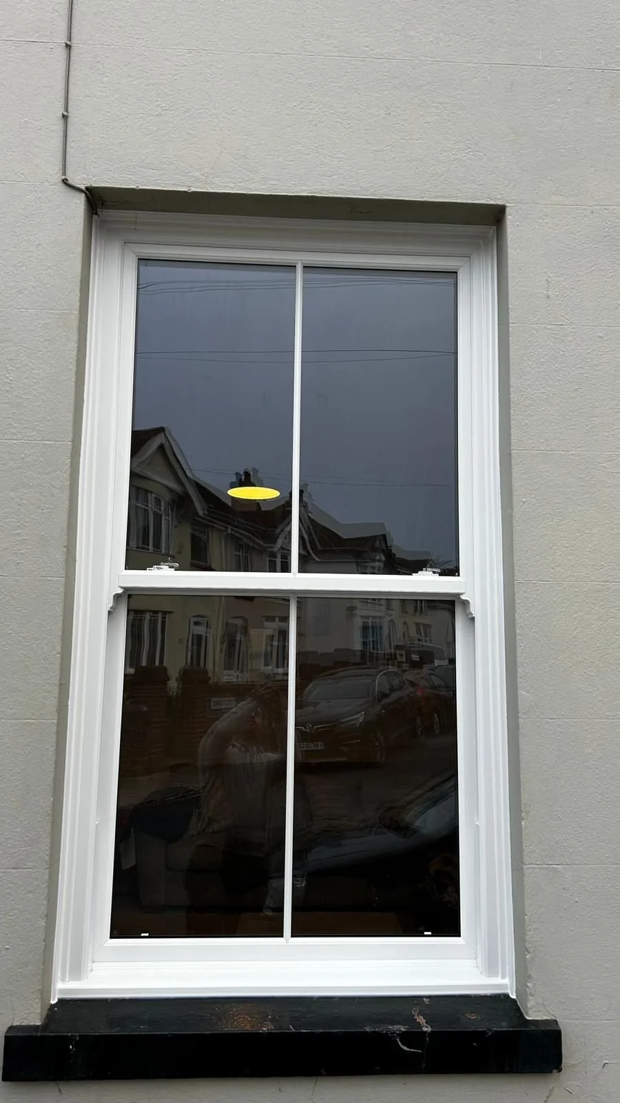 A double-hung window with a white frame on a light gray exterior wall, reflecting houses, cars, and a person taking the photo.