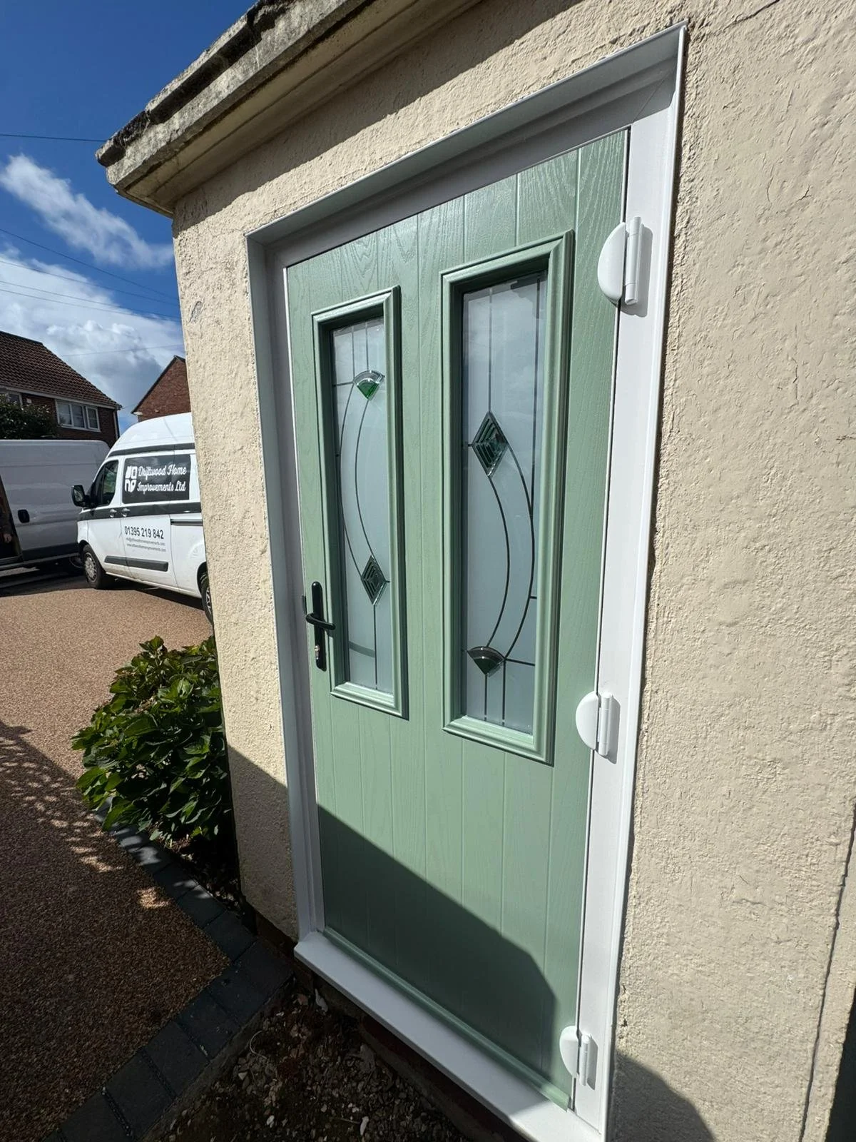 Front door painted light green with decorative glass and black handle, set in a beige textured wall, under a white frame, with a small bush to the left and a driveway with vehicles in the background.