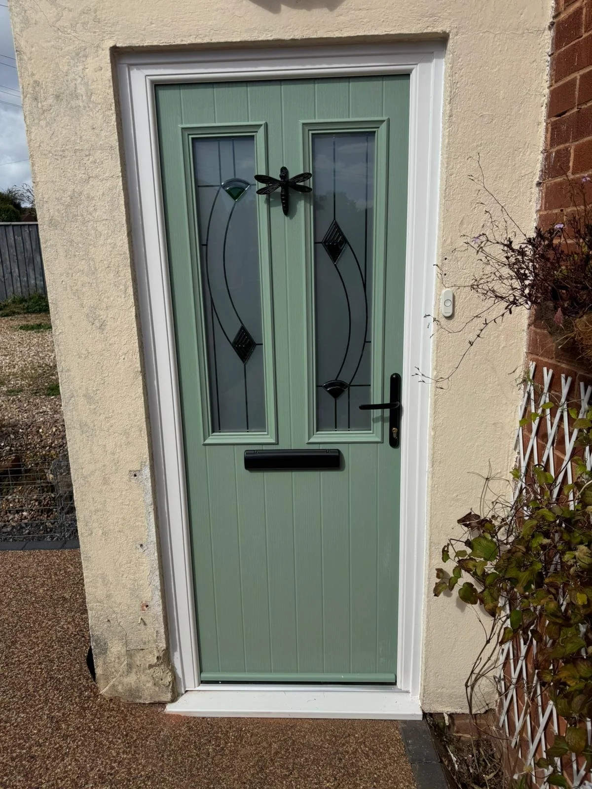 Front door with a black dragonfly decoration, green panels with glass inserts, mail slot, and black handle, set in a cream wall next to a brick wall with a vine.
