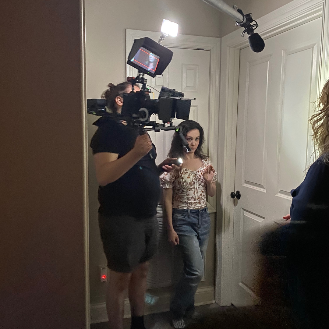Film crew filming a woman in a room with white walls and a closed door, using professional camera equipment and lights.
