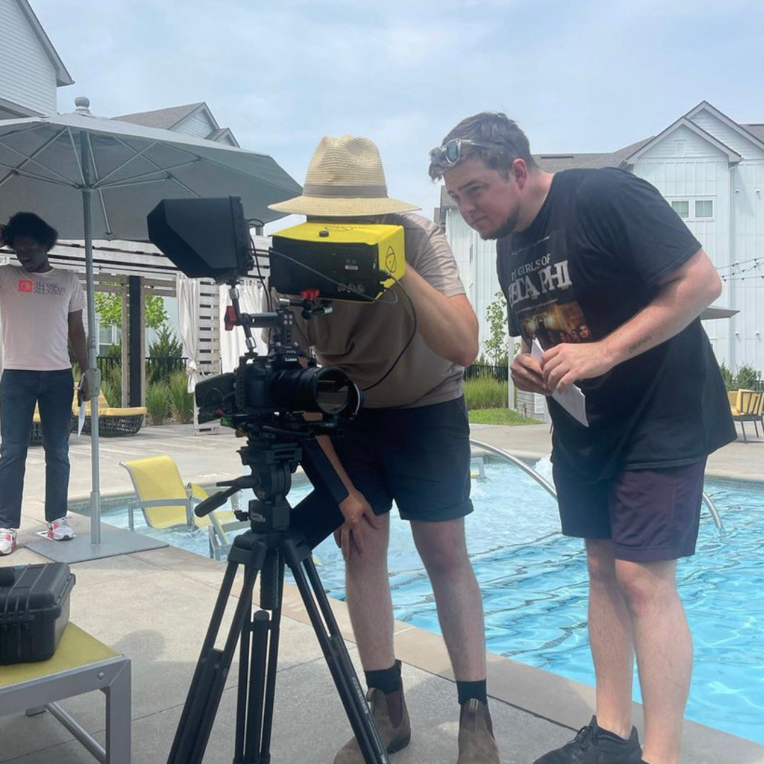 Two men working with a professional film camera outdoors near a swimming pool in a residential area. One man is wearing a straw hat and shorts, crouching near the camera, while the other, wearing a black T-shirt and shorts, is leaning in and holding 