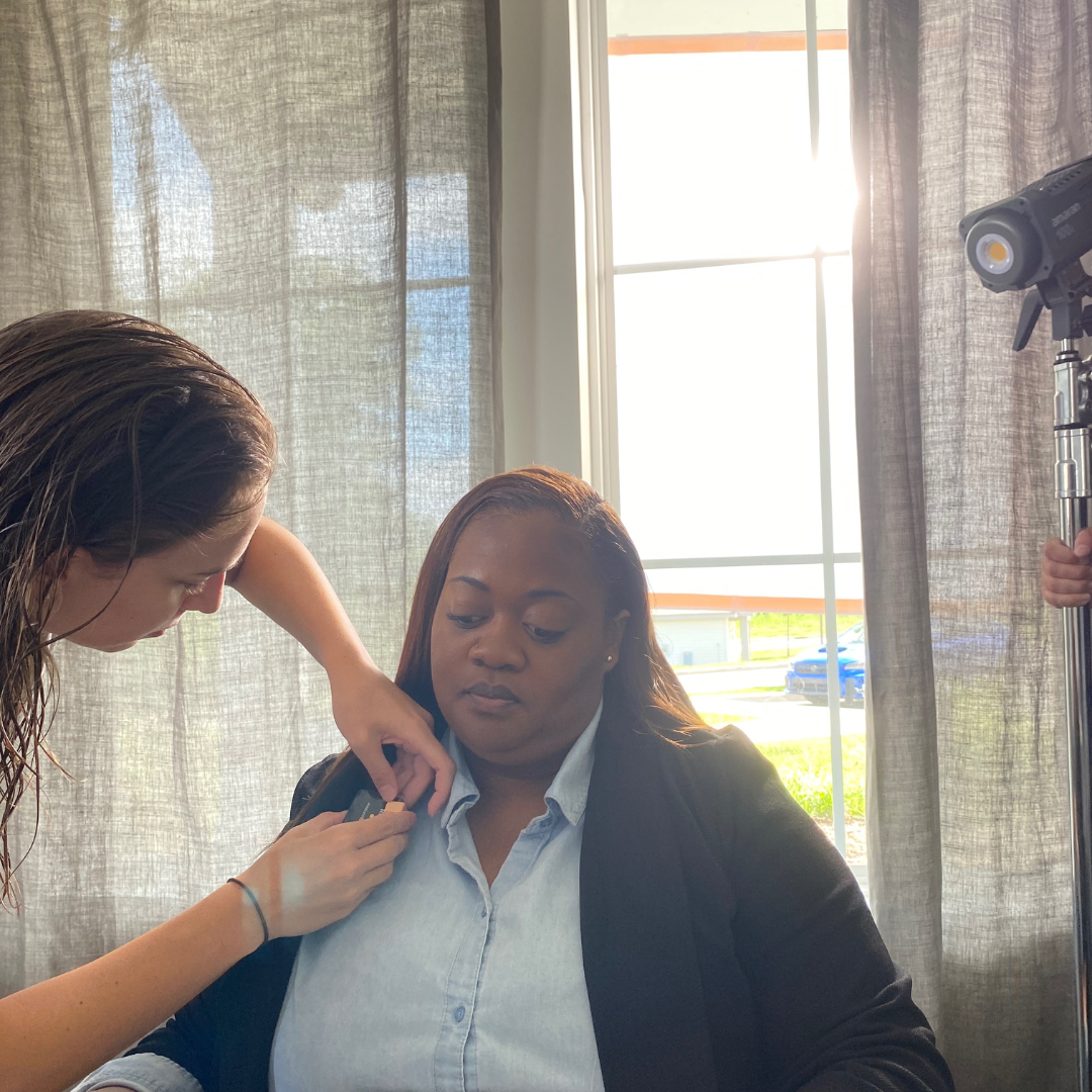 A woman getting her shoulder filmed or photographed indoors with sunlight coming through curtains in the background.