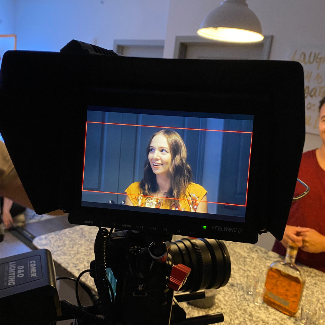 A young woman with long, wavy brown hair is smiling and looking slightly to her right. She is seated in front of a blue panel background, wearing a mustard-colored top with floral embroidery. The photo is being taken through a camera monitor that sho