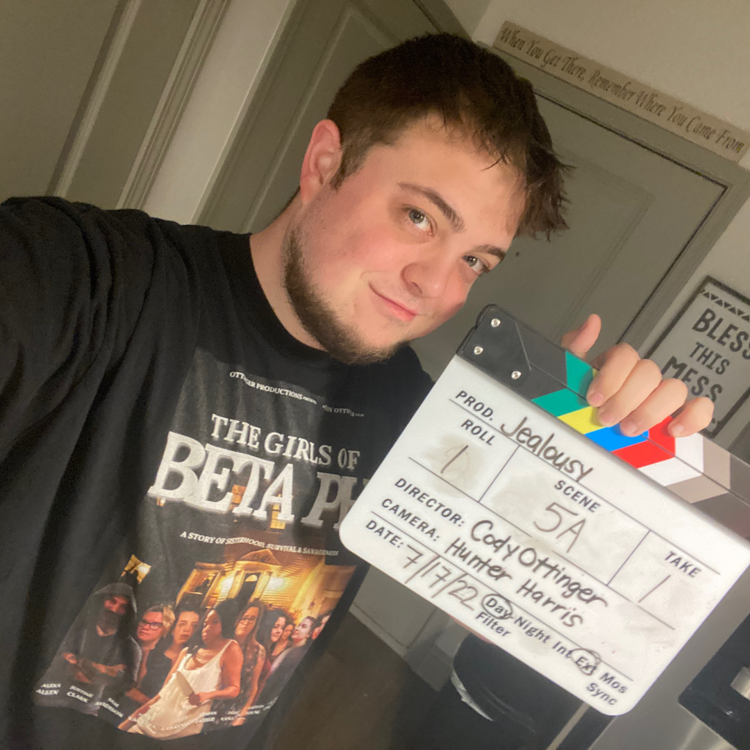 Young man with short brown hair and a beard smiling, holding a movie clapperboard, wearing a black T-shirt with a graphic design featuring women and the text 'The Girls of Beta Pi', standing indoors near a white wall with a framed poster and decorati