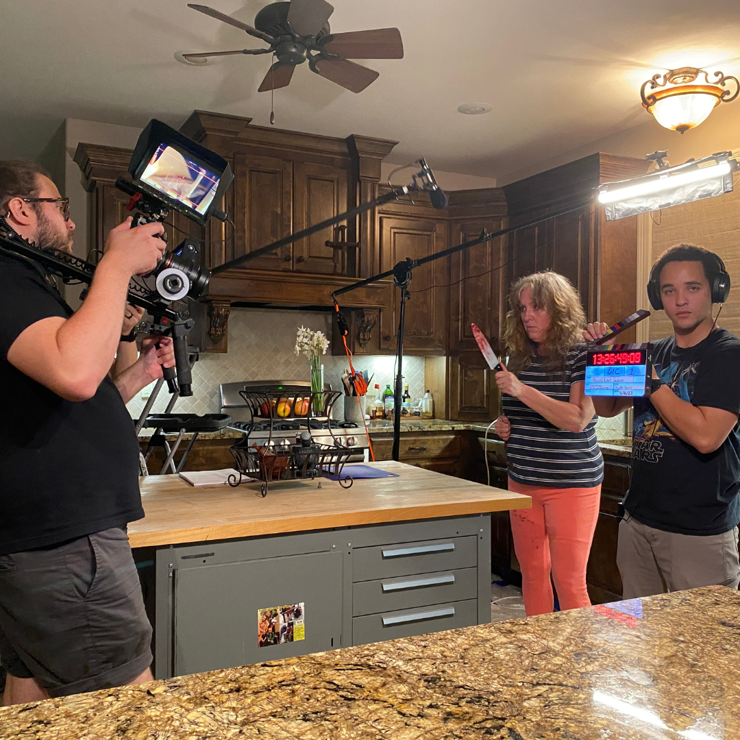Filming kitchen scene with woman holding a knife, crew member with clapperboard, and a cameraman with camera and equipment.
