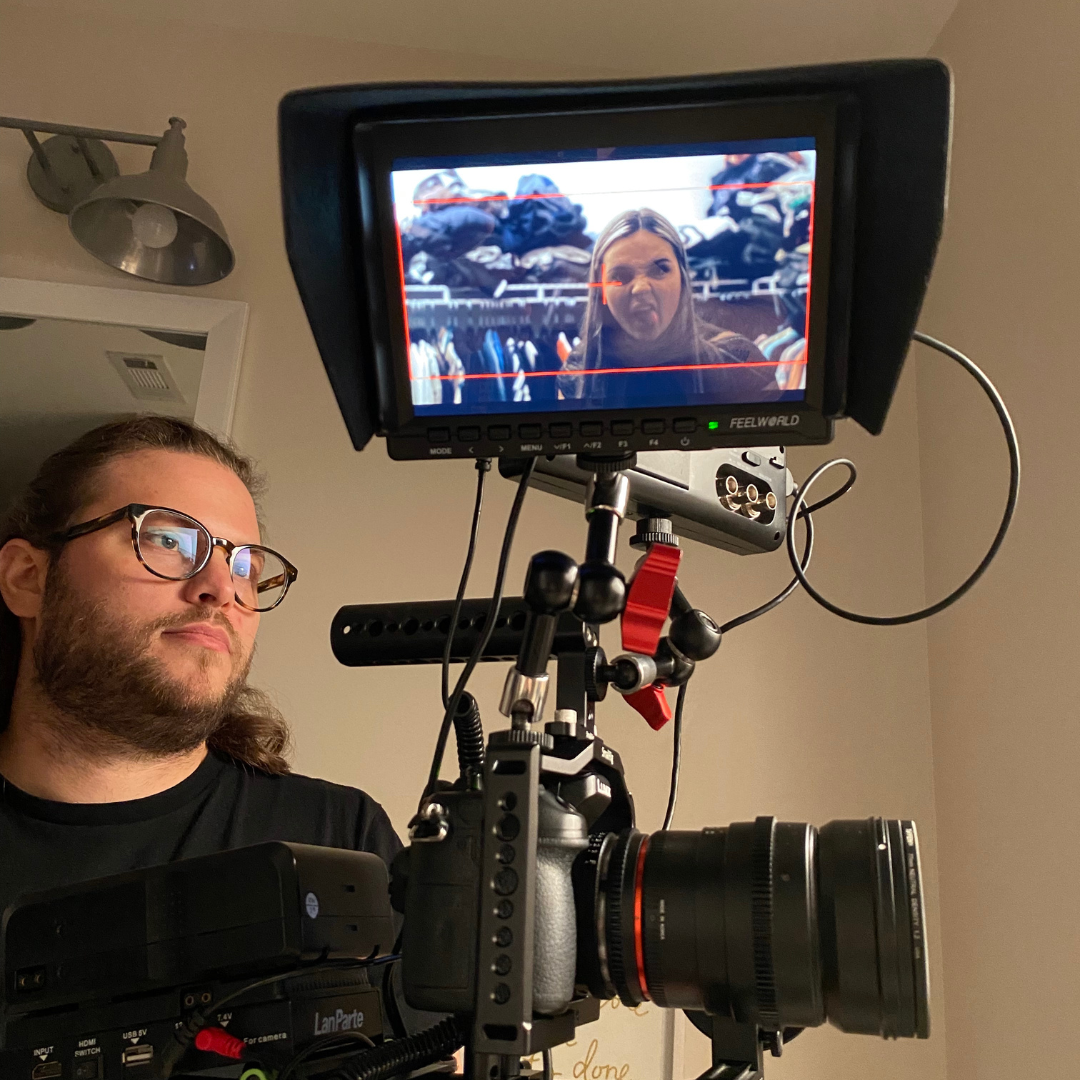 A man with glasses and long hair takes a photo or records a video of a woman making a funny face with her tongue out, seen on a monitor. They are in an indoor setting with a light beige wall and a ceiling light fixture.