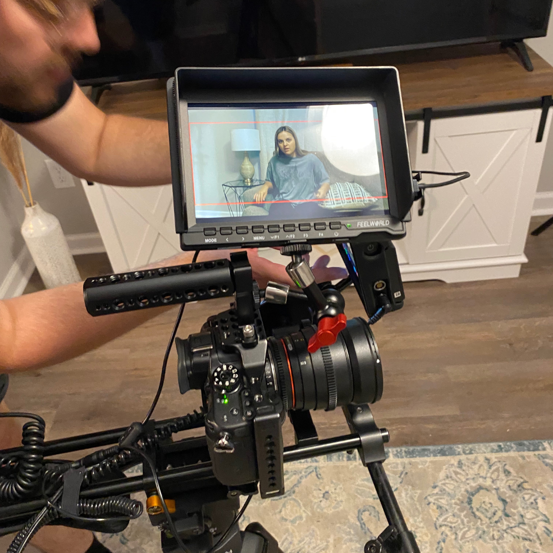 Behind-the-scenes of a video shoot showing a camera operator filming a woman sitting on a bed in a bedroom, with a monitor displaying her image.
