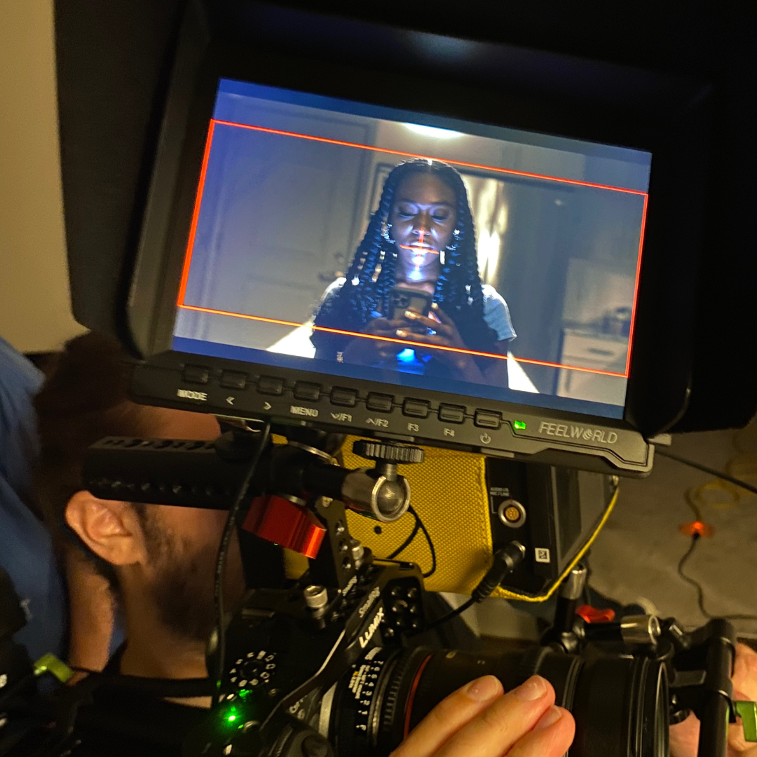 A woman with braided hair is looking at a smartphone, visible on the monitor in front of her.