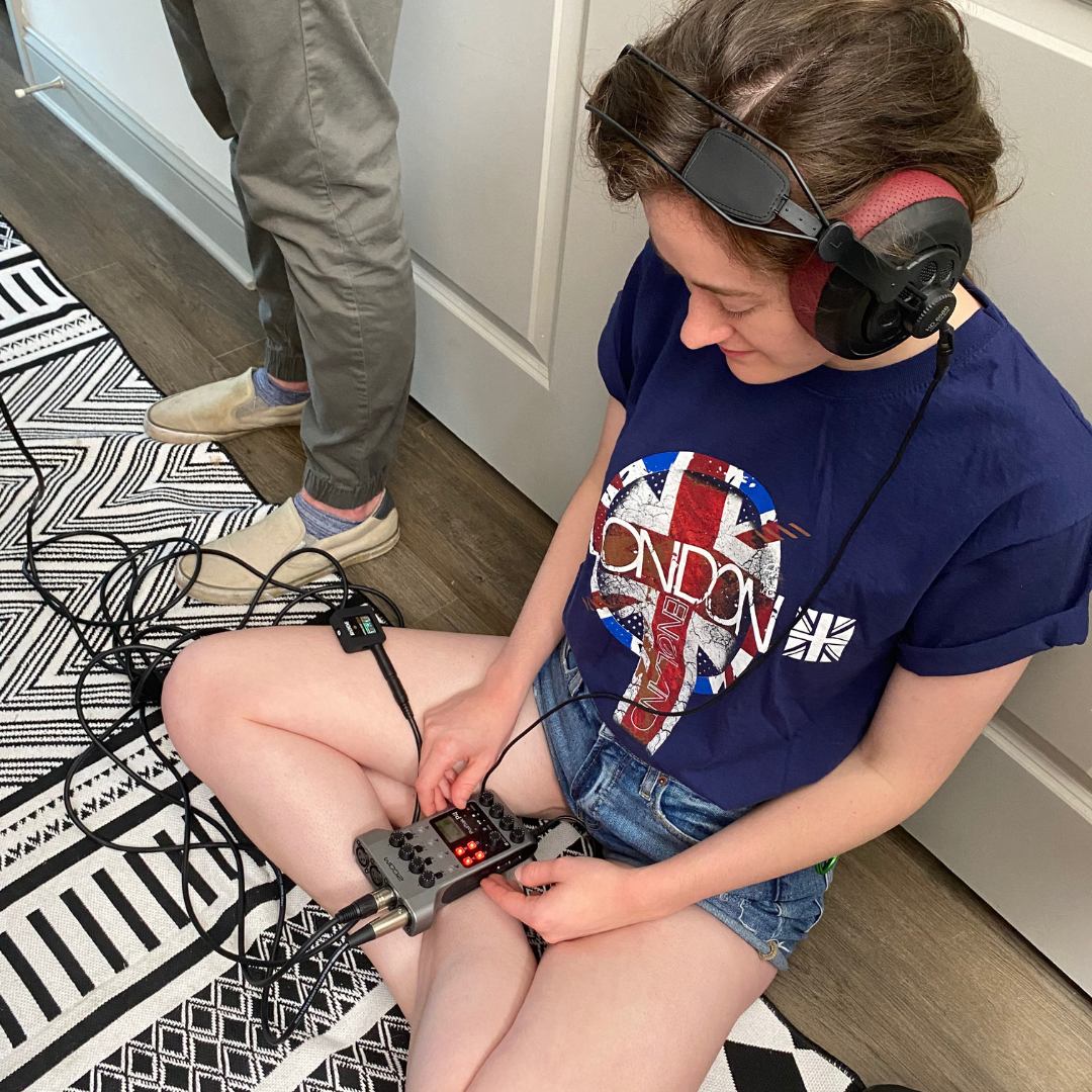 A young woman sitting on the floor with headphones, operating an audio mixer with multiple cables, while another person stands nearby. The woman wears a dark blue T-shirt with a graphic and denim shorts, and the floor has a black and white patterned 