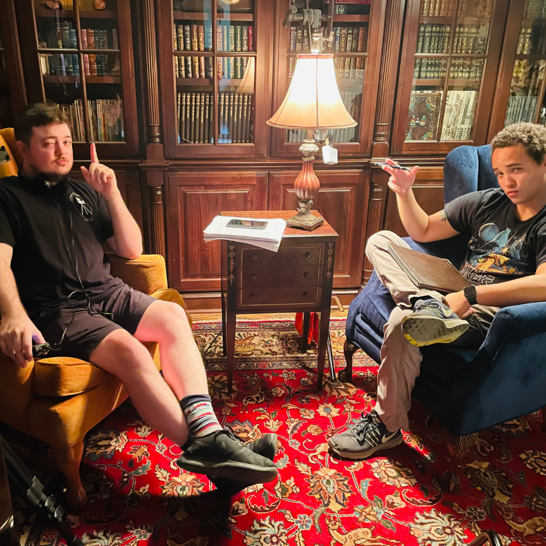 Two young men seated on chairs in a cozy room with wooden bookshelves, a small wooden table, a table lamp, and a patterned red area rug. One is sitting on a mustard armchair, raising his index finger, and the other is seated on a blue armchair, holdi