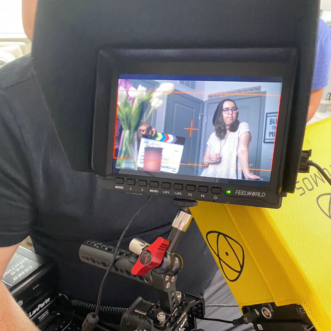 A woman stands in front of a gray door, wearing glasses and a white top, holding a drink, during a filming or photo shoot, with a clapperboard and flowers nearby.