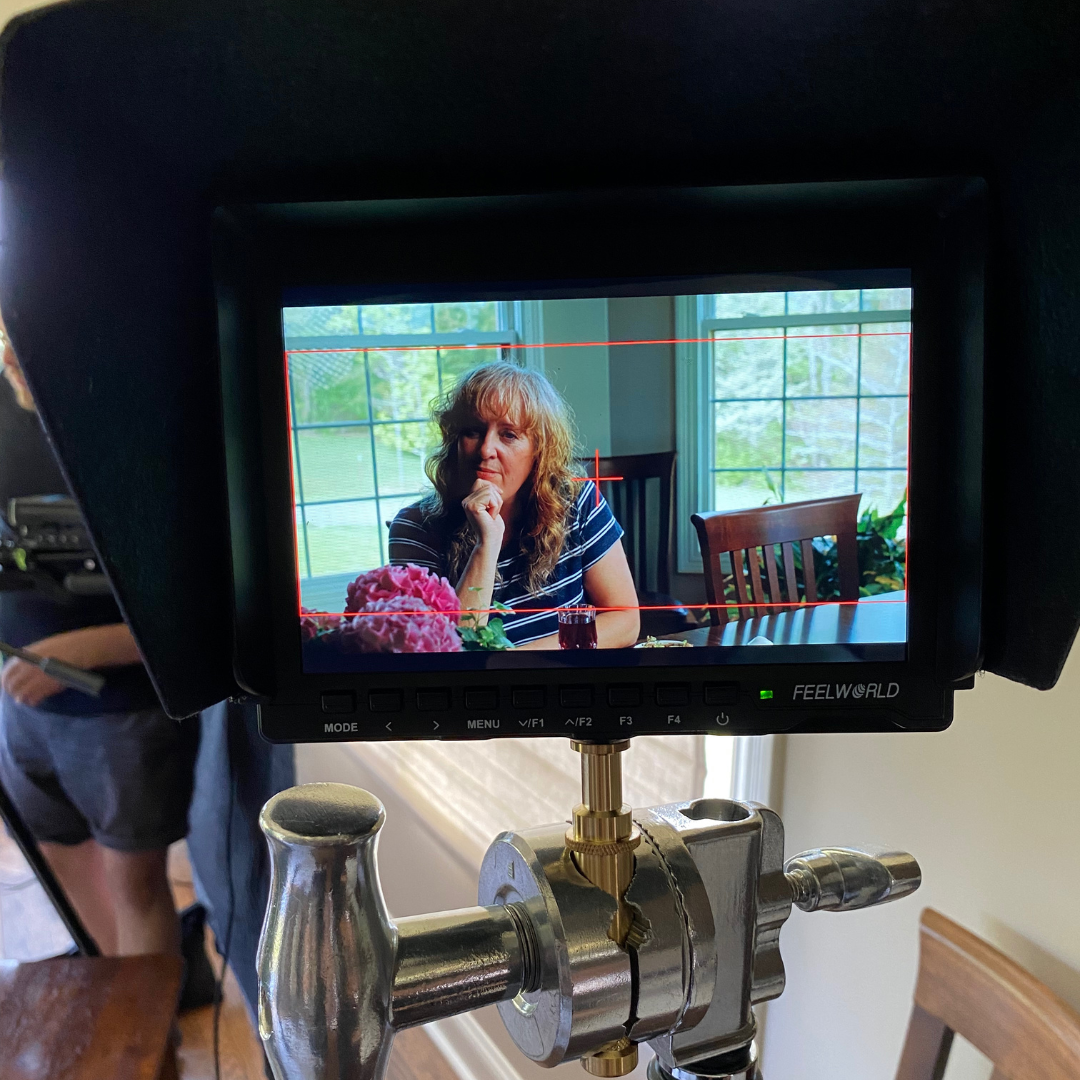 A camera monitor shows a woman with curly hair and a striped shirt sitting at a dining table near windows with green outdoor scenery, with pink flowers and a glass of dark-colored beverage on the table.