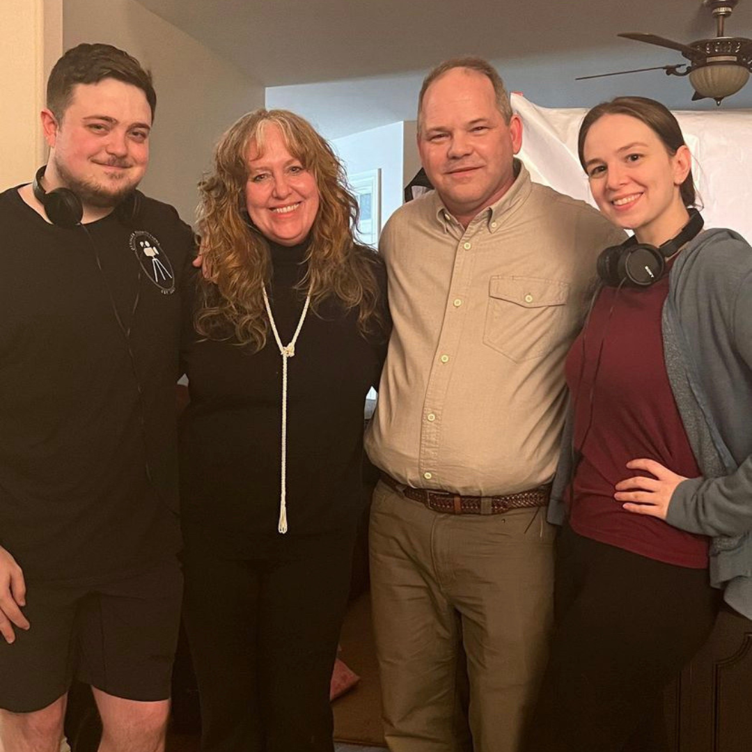 A group of four people standing close together indoors, smiling at the camera. Two young women, one on each side, are wearing headphones around their necks. The middle two individuals are an older woman with curly hair and a man in a beige button-up 