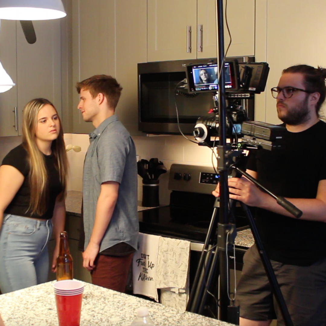Behind the scenes of a video shoot in a kitchen with a man operating a camera on a tripod, a woman and a man standing close together, and a cameraman filming the scene.